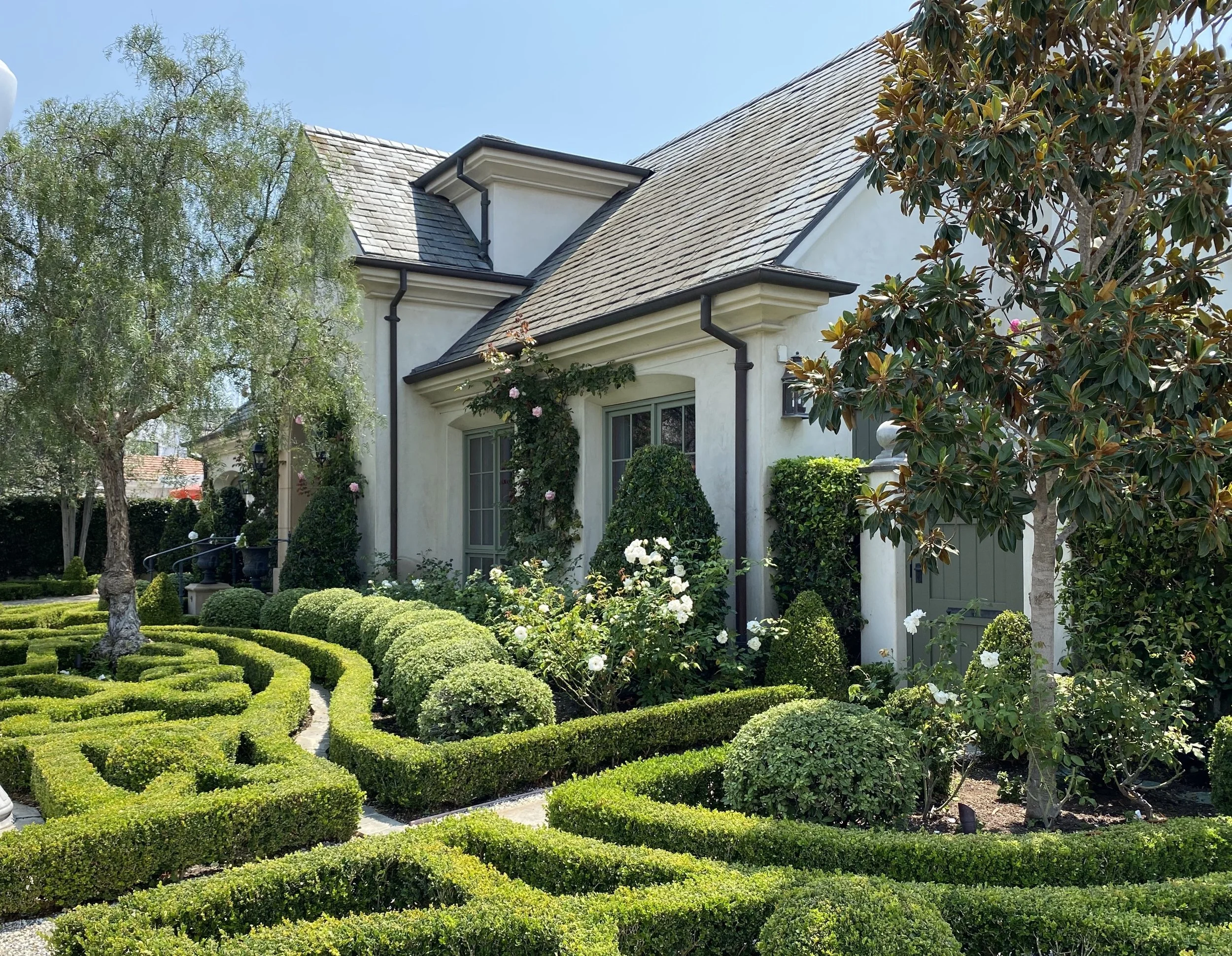 front yard of a house with neatly trimmed hedges and bushes, flowering plants, trees, and a white house with a sloped roof and black gutters