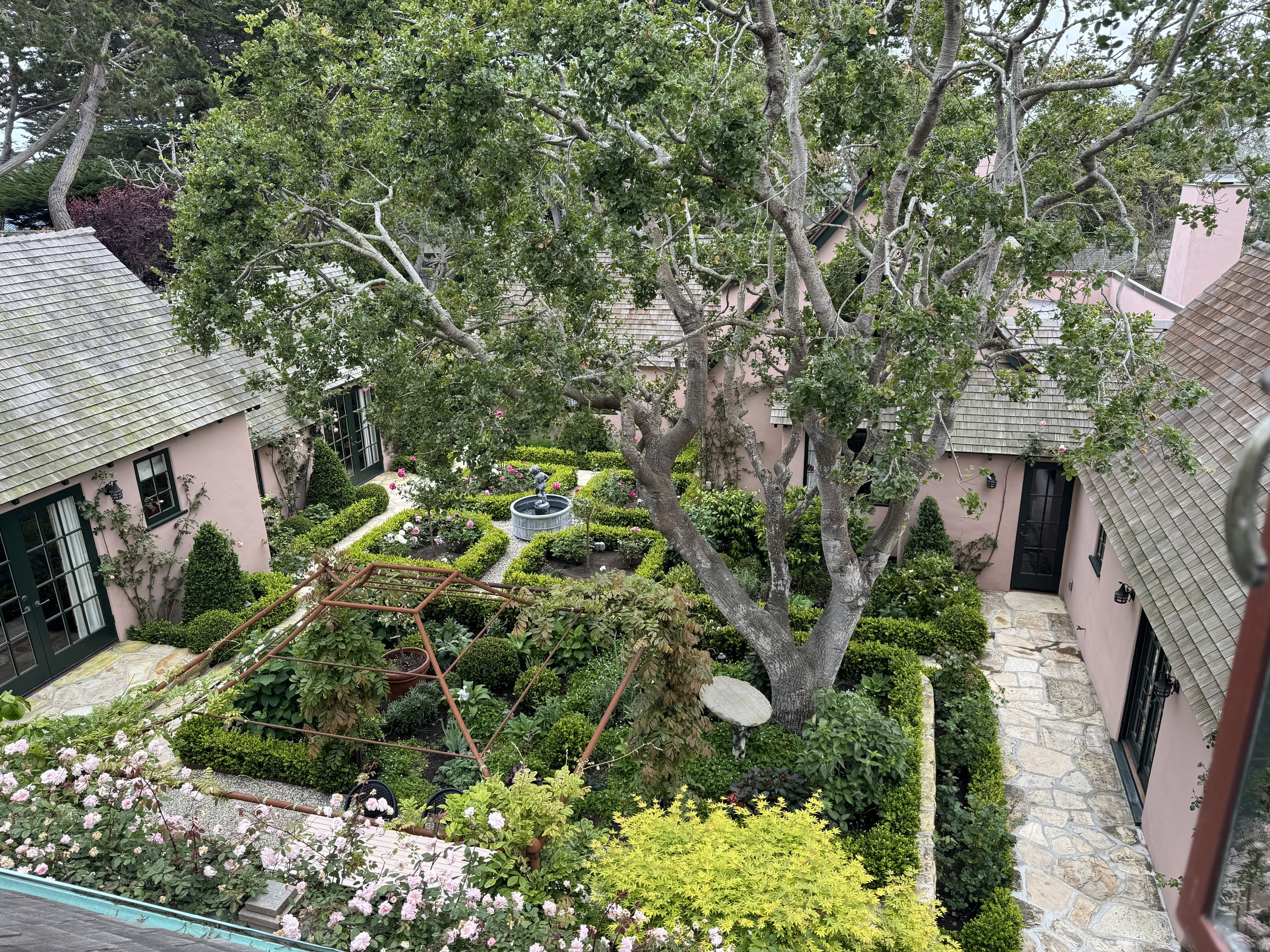 A lush garden courtyard surrounded by pink buildings with gray tiled roofs. The garden features a large tree, trimmed bushes, flowering plants, a stone pathway, and a small fountain.