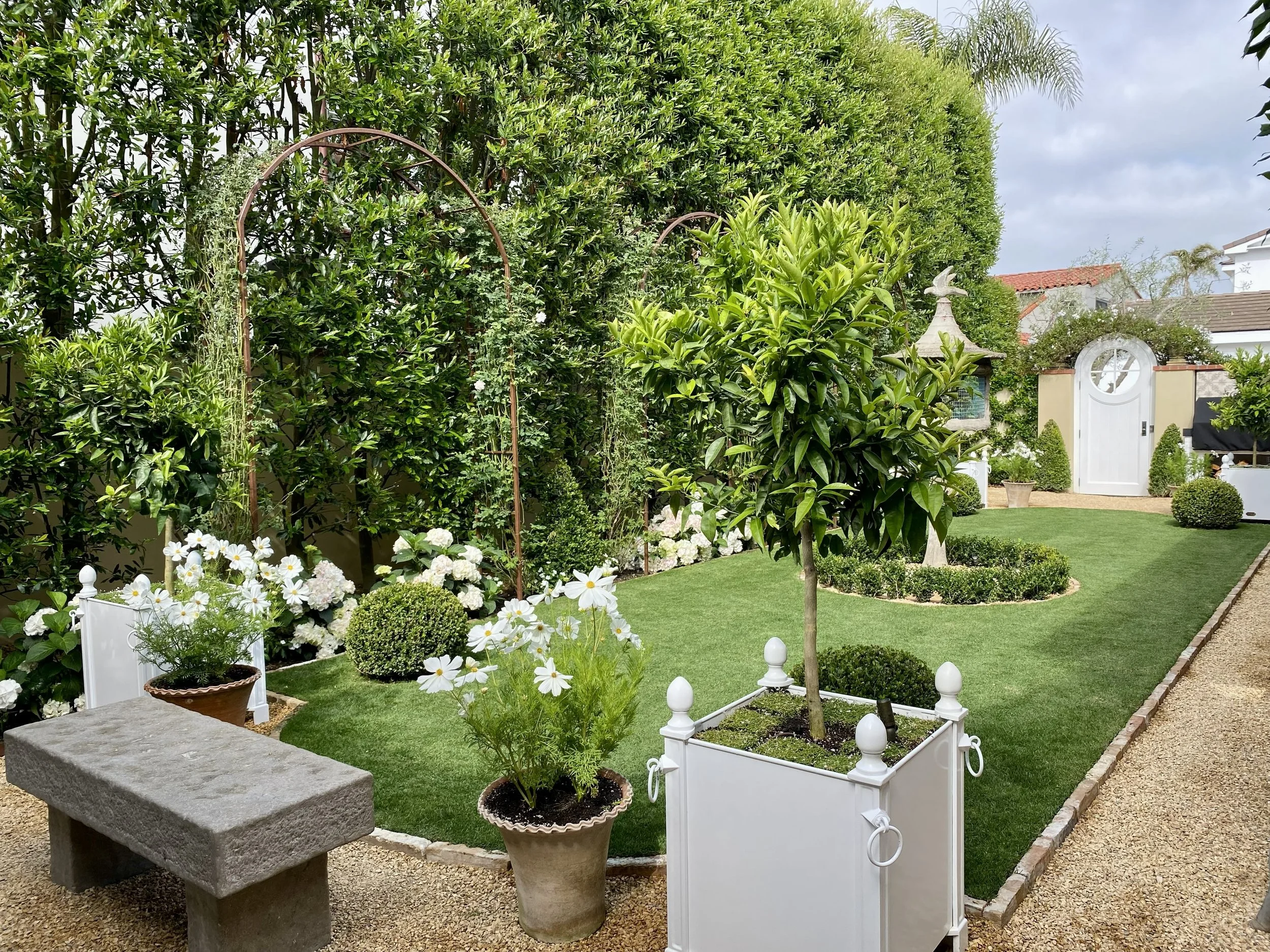 A well-maintained garden with green grass, potted plants, flowering bushes, trees, a stone bench, and decorative garden structures including a gated arch and a small white building in the background.