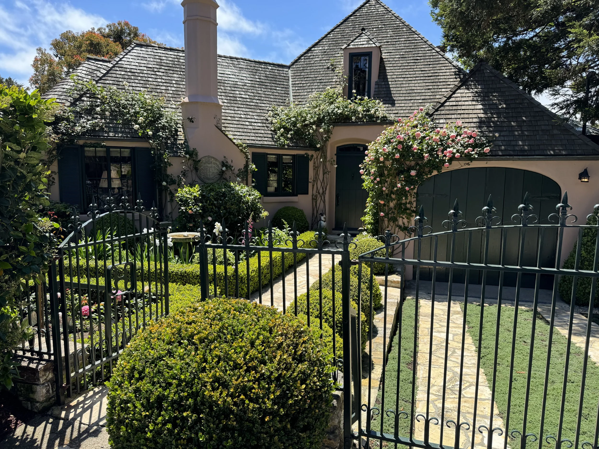 A charming house with a slate roof, surrounded by a garden with manicured bushes and flowering vines, enclosed by a black wrought-iron gate.