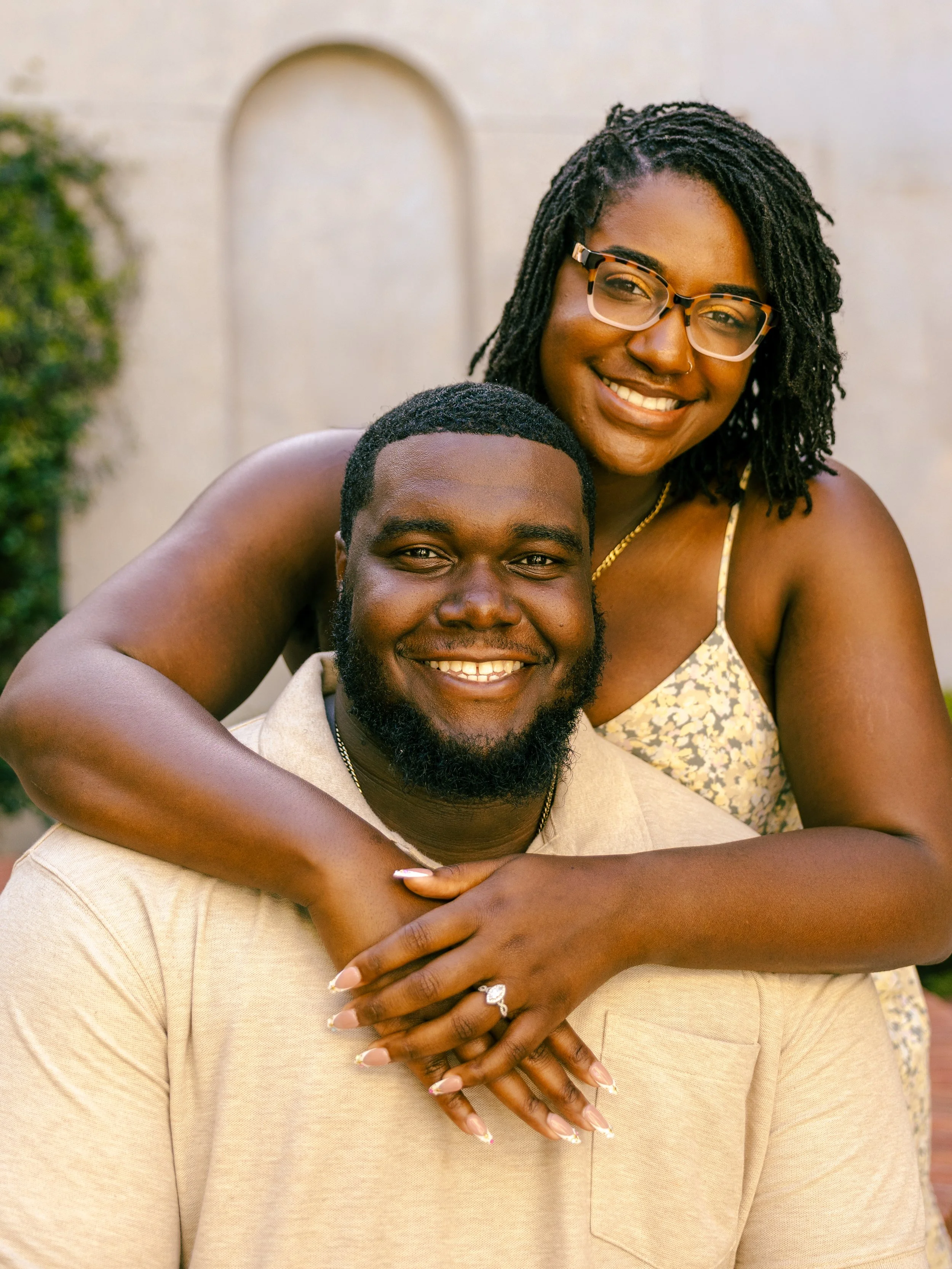 A happy couple, a woman with dreadlocks and glasses smiling and embracing a man with a beard and short hair, both smiling at the camera outdoors.