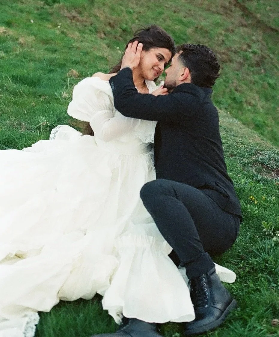 A couple sitting on grass, sharing an intimate moment, with the woman in a white dress and the man in a black outfit, holding each other's faces.