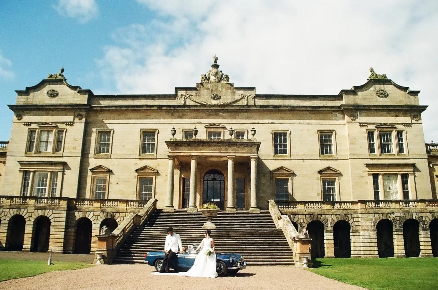 A large, historic mansion with stone steps leading to the entrance, featuring six large windows, decorative elements, and statues on top. A man and woman in wedding attire are standing near a vintage blue convertible car parked in front.