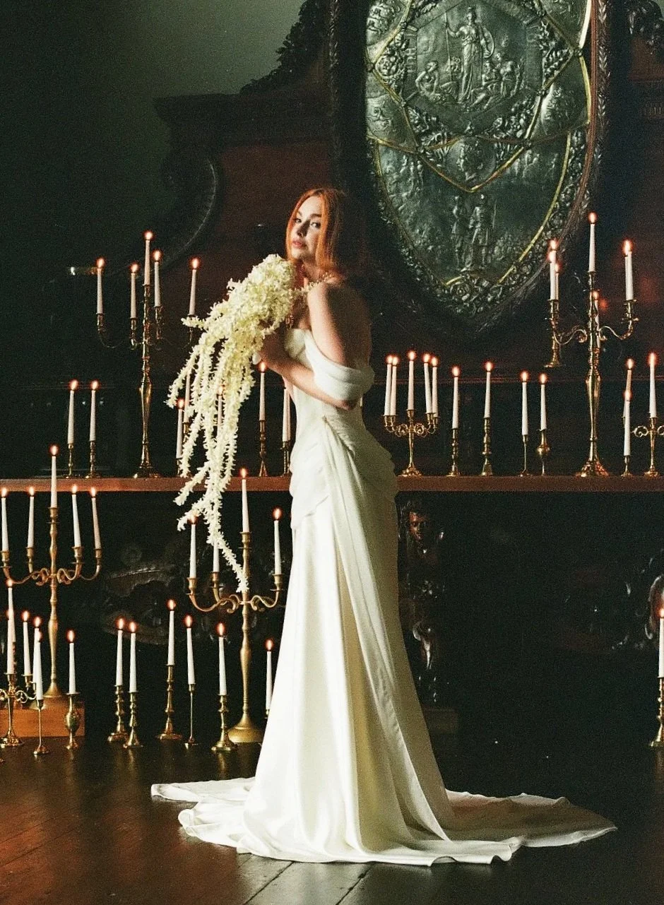 A woman in a white gown holding a large bouquet of white flowers, standing in a dimly lit room surrounded by lit candelabras and an ornate, metallic artwork behind her.