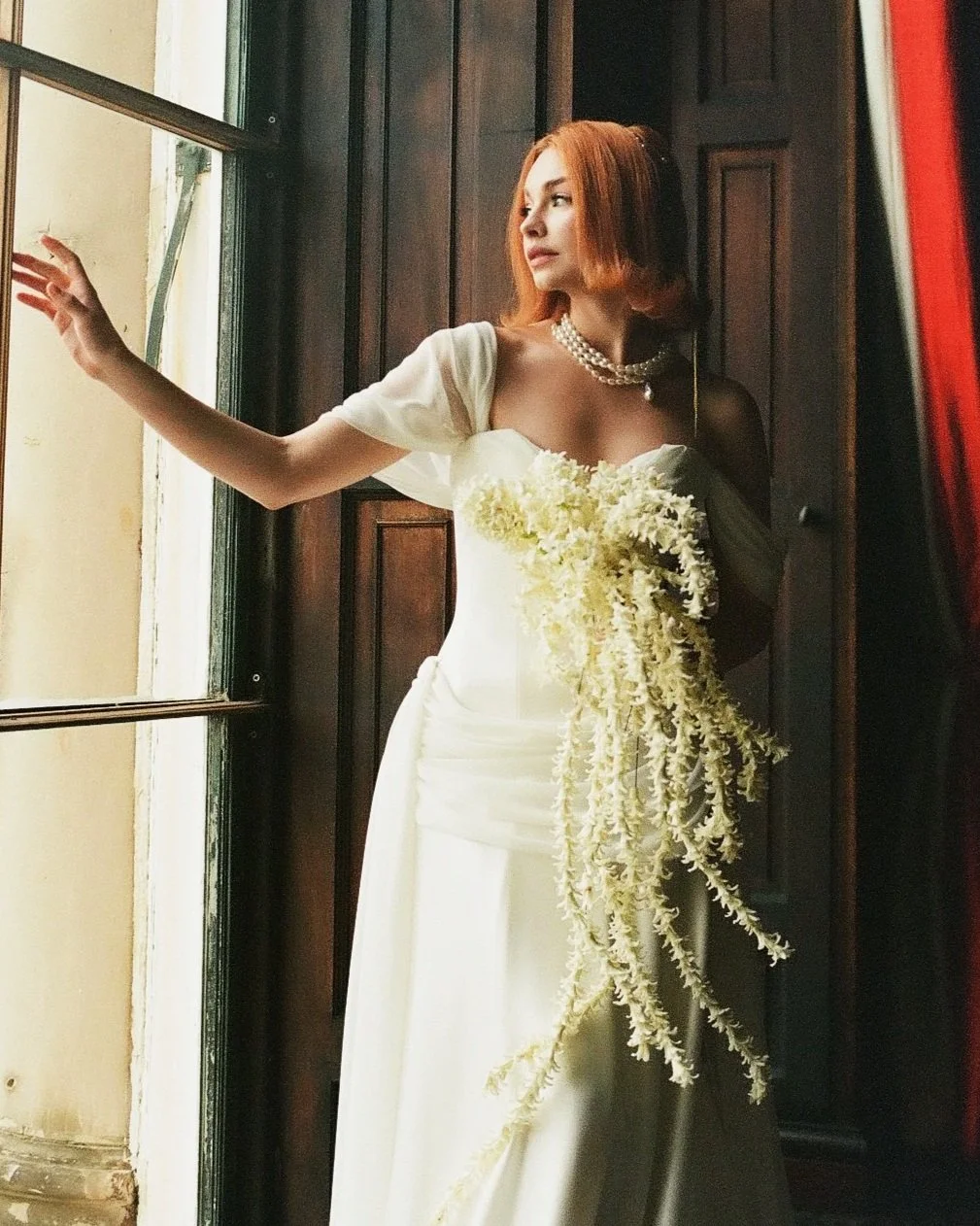 A woman in a white wedding dress holding a cascading bouquet of white flowers, standing near a window with natural light.