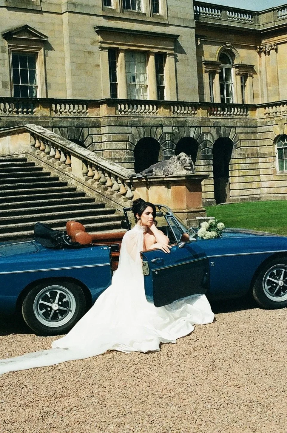 A woman in a white wedding dress sitting on the door of a blue vintage convertible car, parked in front of a large, historic stone mansion with stairs and lions statues
