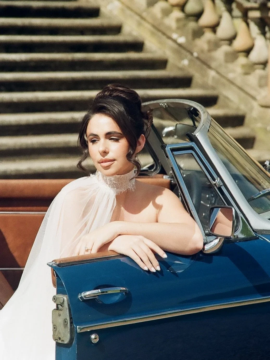A woman with dark hair styled in an updo, wearing a white dress with lace details, leaning on the door of a vintage blue convertible car parked outdoors, with stone stairs in the background.