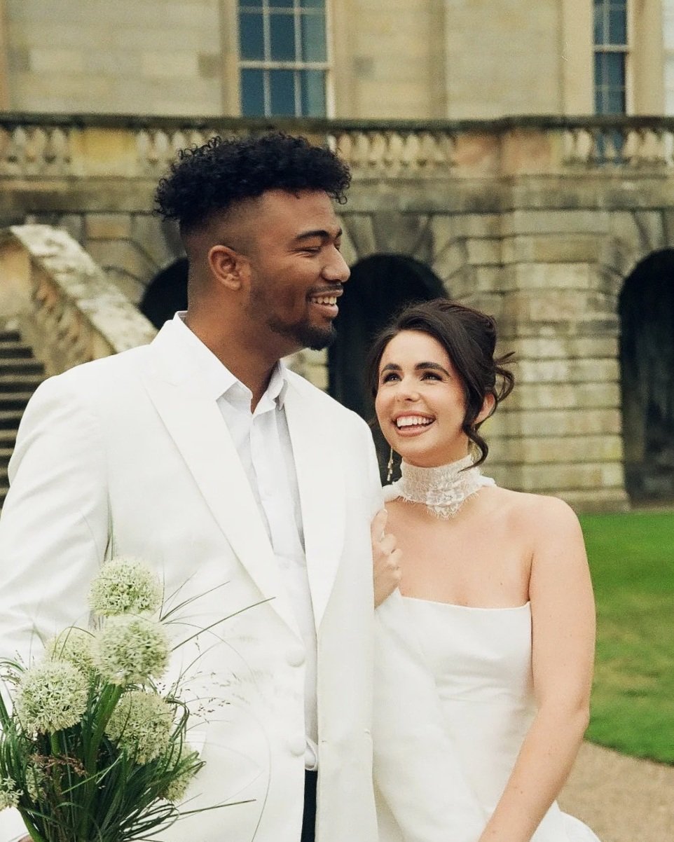 A newlywed couple dressed in wedding attire, smiling, standing outdoors in front of a stone building with stairs and arches.