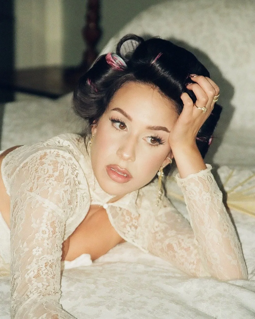 Woman with black hair styled with pink rollers, lying on a bed wearing a white lace top, resting her head on her hand, with a thoughtful expression.