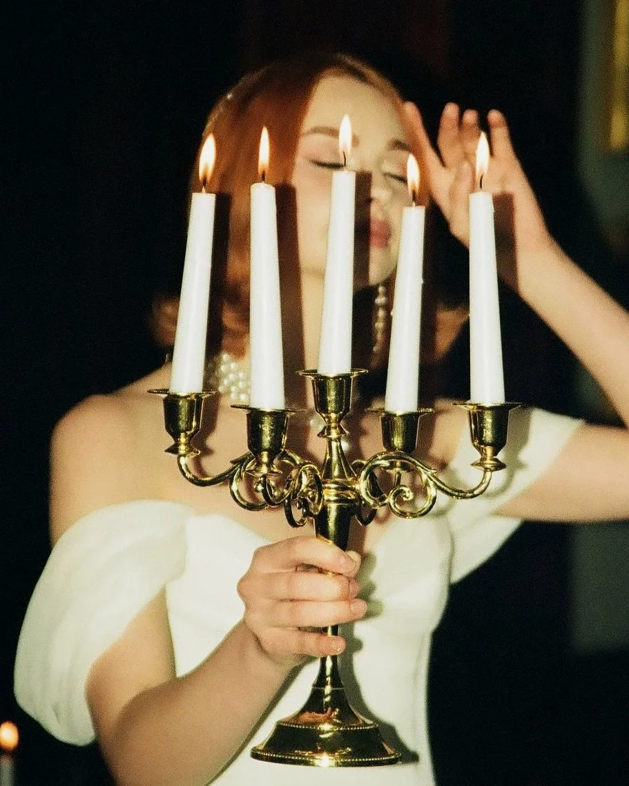 Young woman in an elegant white dress holding a lit candelabrum with five candles, with a dark background.