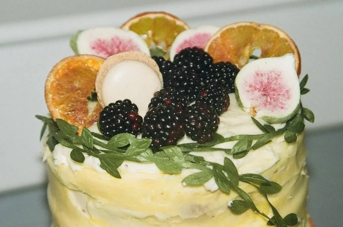 A layered cake topped with sliced figs, dried orange slices, blackberries, and greenery.