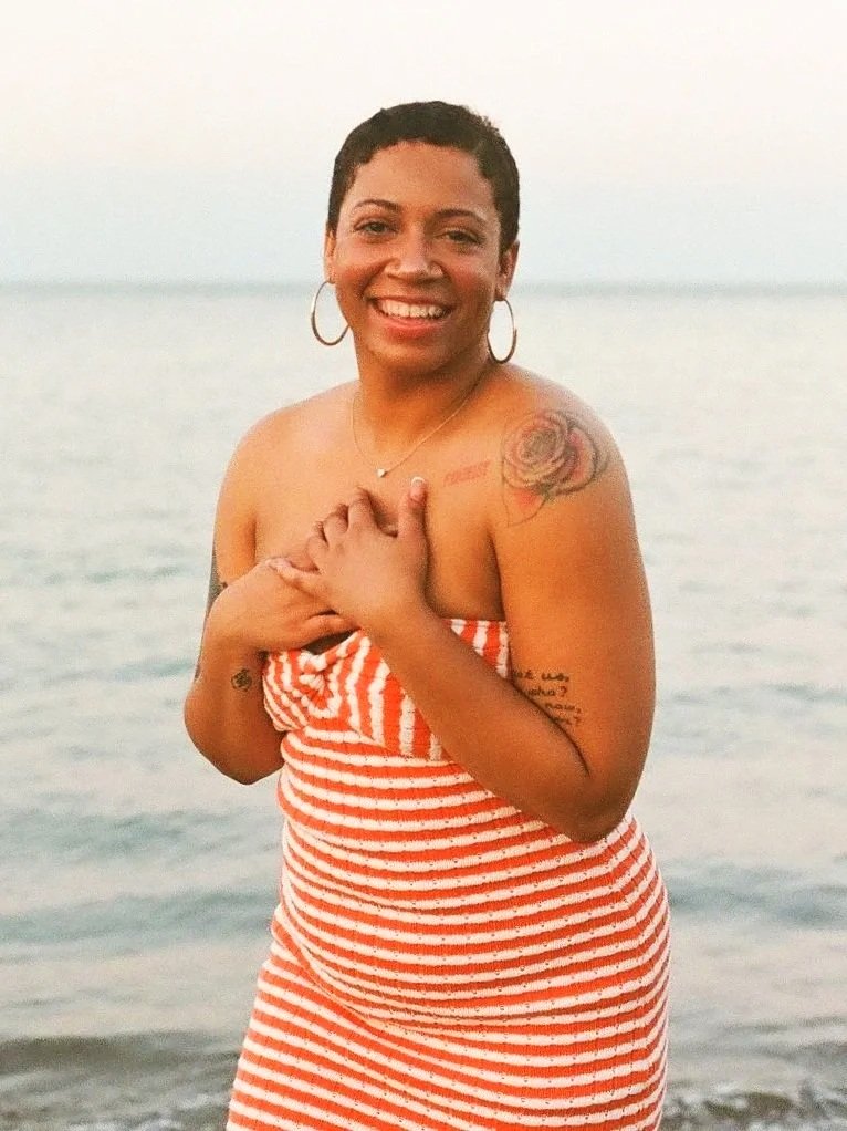 Smiling woman standing on a beach, wearing a red and white striped towel dress, with tattoos on her arm and shoulder, earrings, and a necklace.