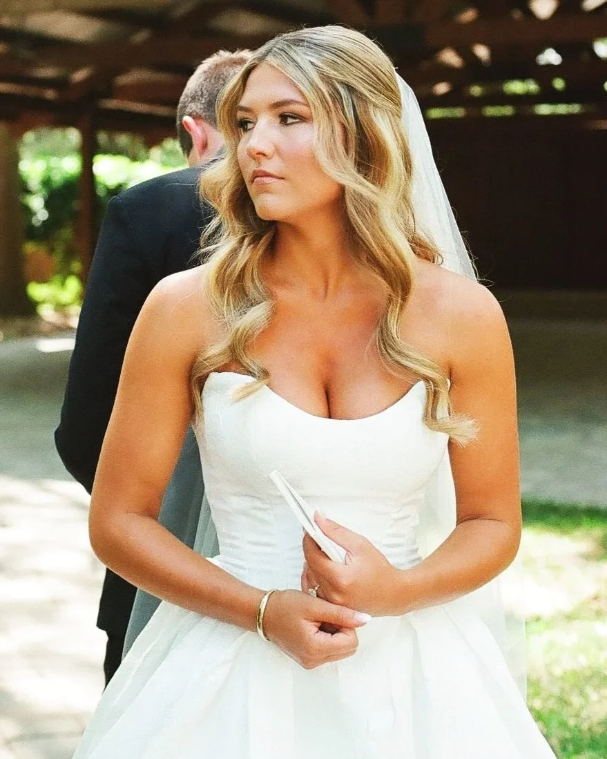A woman in a wedding dress holding a small white object, standing outdoors with a man in a black suit behind her, partially obscured.