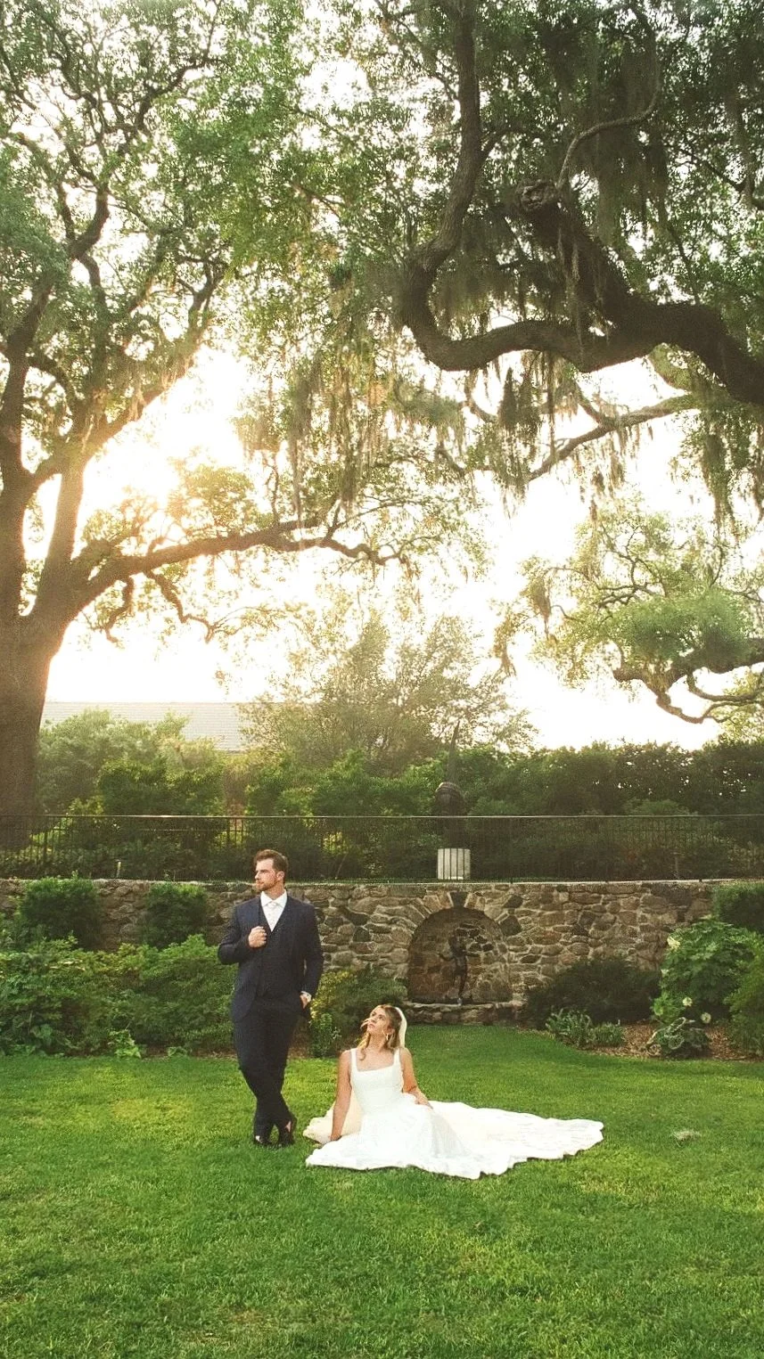 A bride in a white wedding dress sitting on grass and a groom in a suit standing beside her in a lush garden with large trees and stone retaining wall.