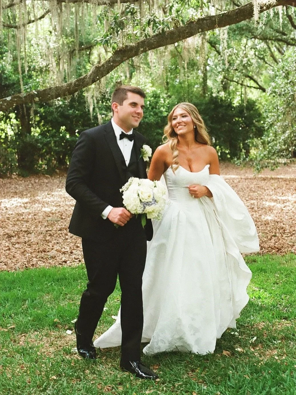 A bride and groom standing outdoors in a wooded area, the groom in a black tuxedo with a bow tie holding a bouquet of white flowers, and the bride in a strapless white wedding gown holding her dress.