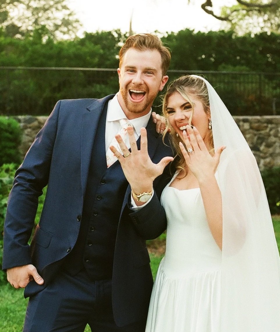 A newlywed couple smiling and showing off their wedding rings outdoors during daytime, with trees and a stone wall in the background.