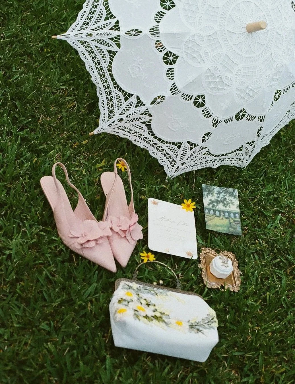 Collection of wedding or bridal accessories on grass, including a white lace umbrella, pink shoes with flower decoration, a wedding invitation, a small floral framed picture, and a white box with floral design.