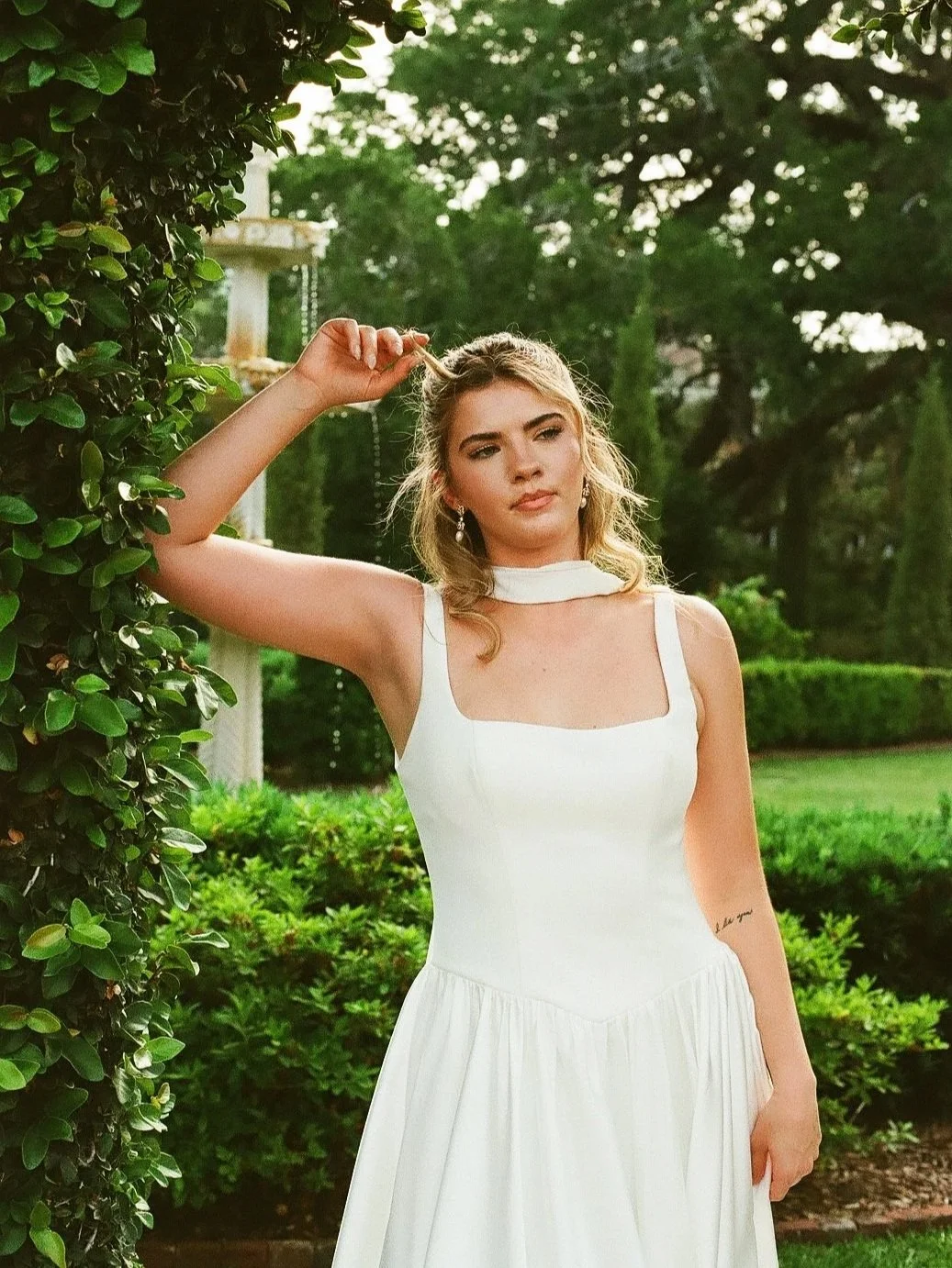 A woman in a white dress standing outdoors near bushes and trees, with one arm raised touching her hair, looking slightly away from the camera.