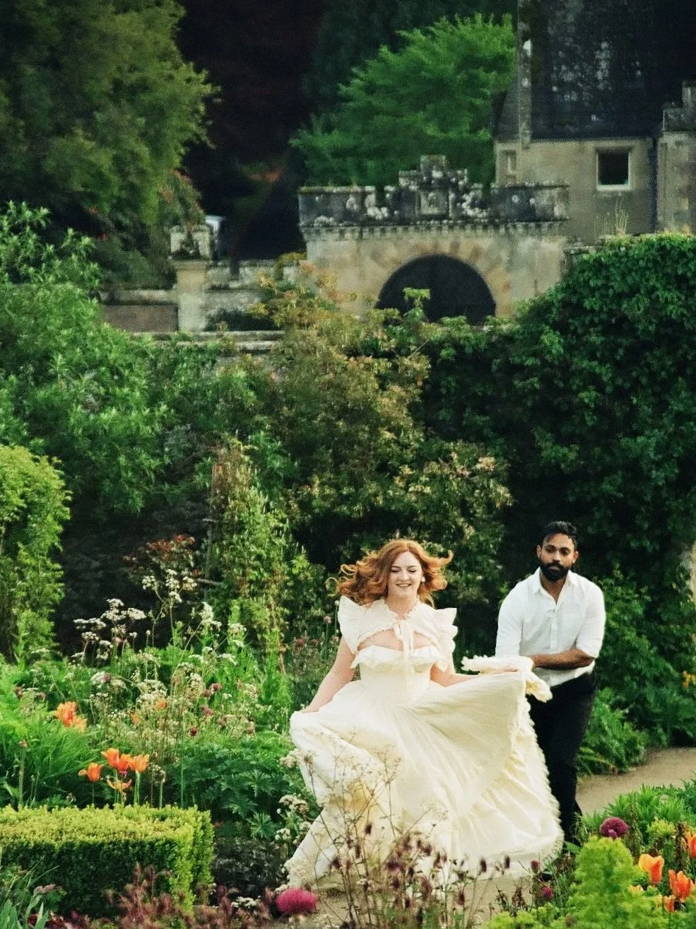 A woman in a flowing cream dress running through a lush garden with a man following her, surrounded by colorful flowers and green foliage.