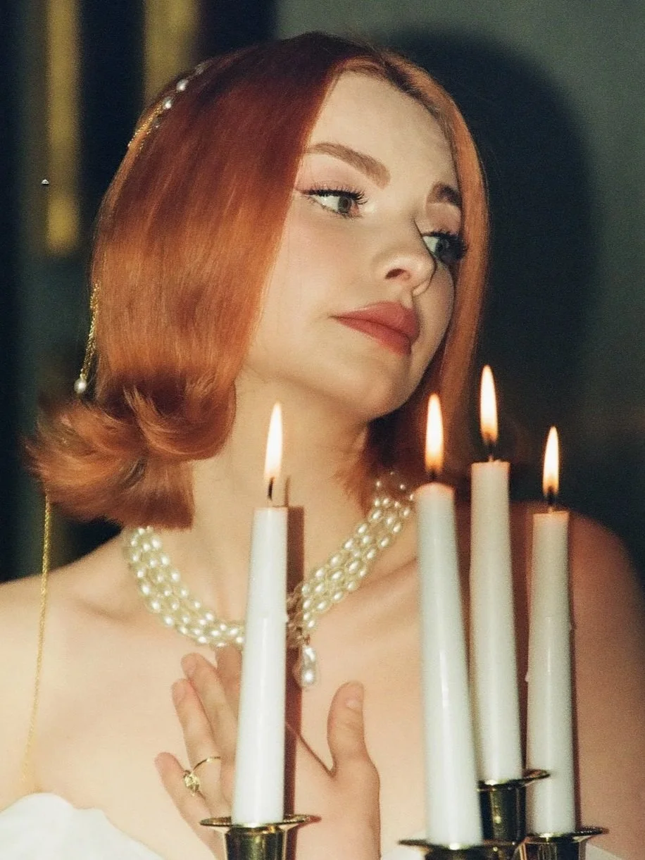 A woman with red hair and pearl jewelry, standing near lit white candles.