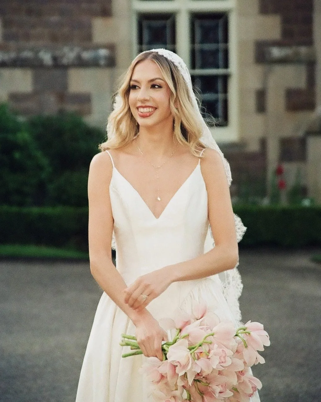 A smiling woman in a wedding dress holding a large bouquet of pink flowers outdoors, with a stone building in the background.