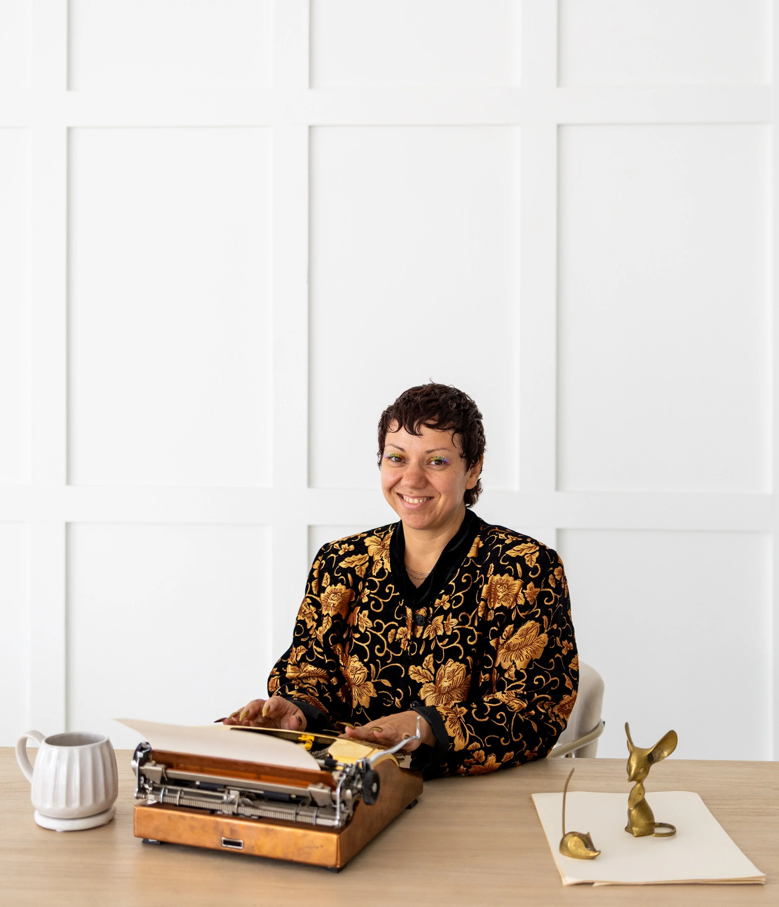 A woman with short curly hair and colorful eye makeup, wearing a black and gold floral patterned blazer, sitting at a light wood table with a vintage typewriter, a white mug, a gold rabbit figurine, a gold crescent moon sculpture, and a sheet of pape