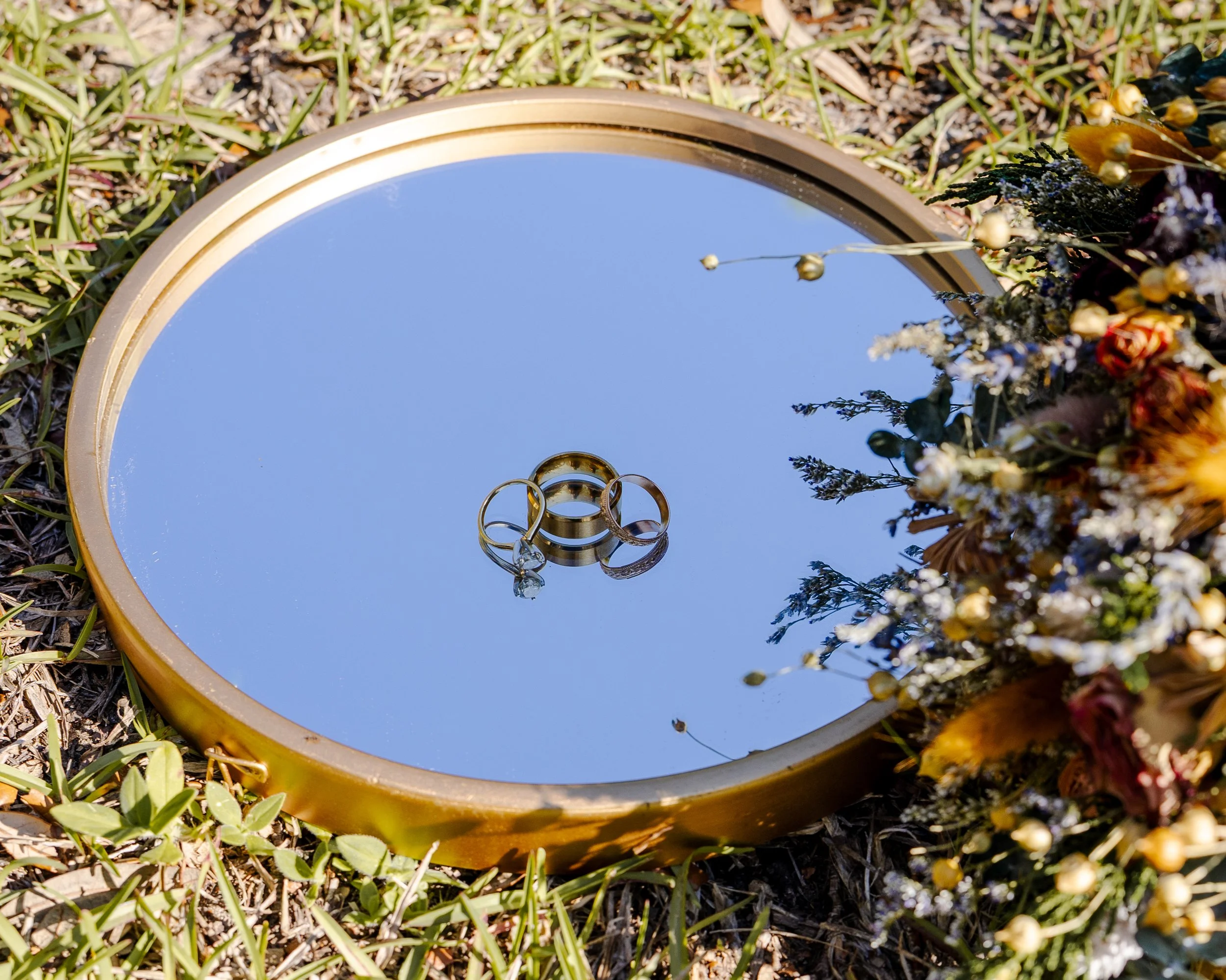 A round mirror with a golden rim lying on grass and surrounded by flowers and greenery, reflecting the blue sky.