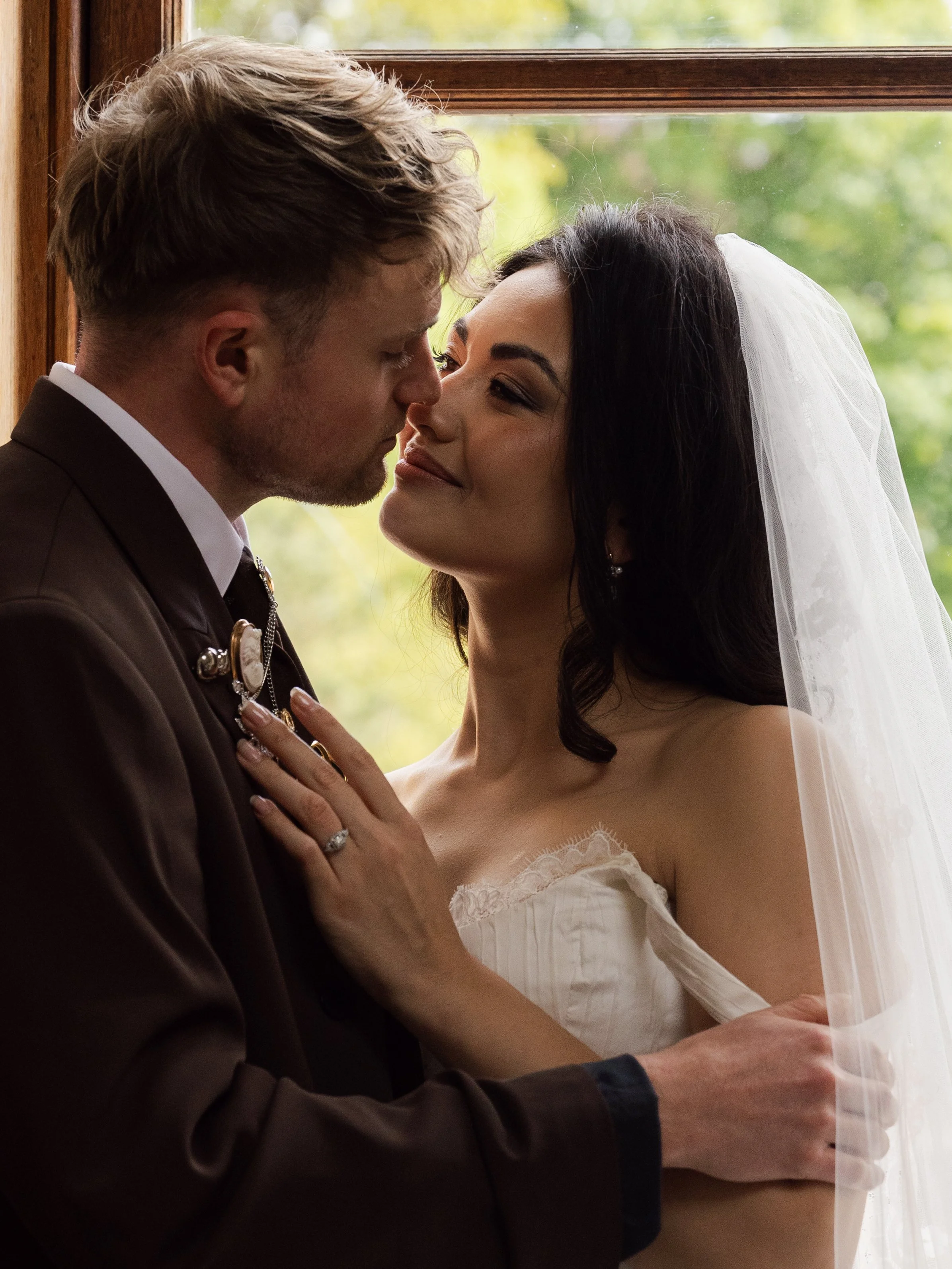 A bride and groom standing close together inside near a window, with the groom in formal attire and the bride in a wedding dress and veil, gazing affectionately at each other.