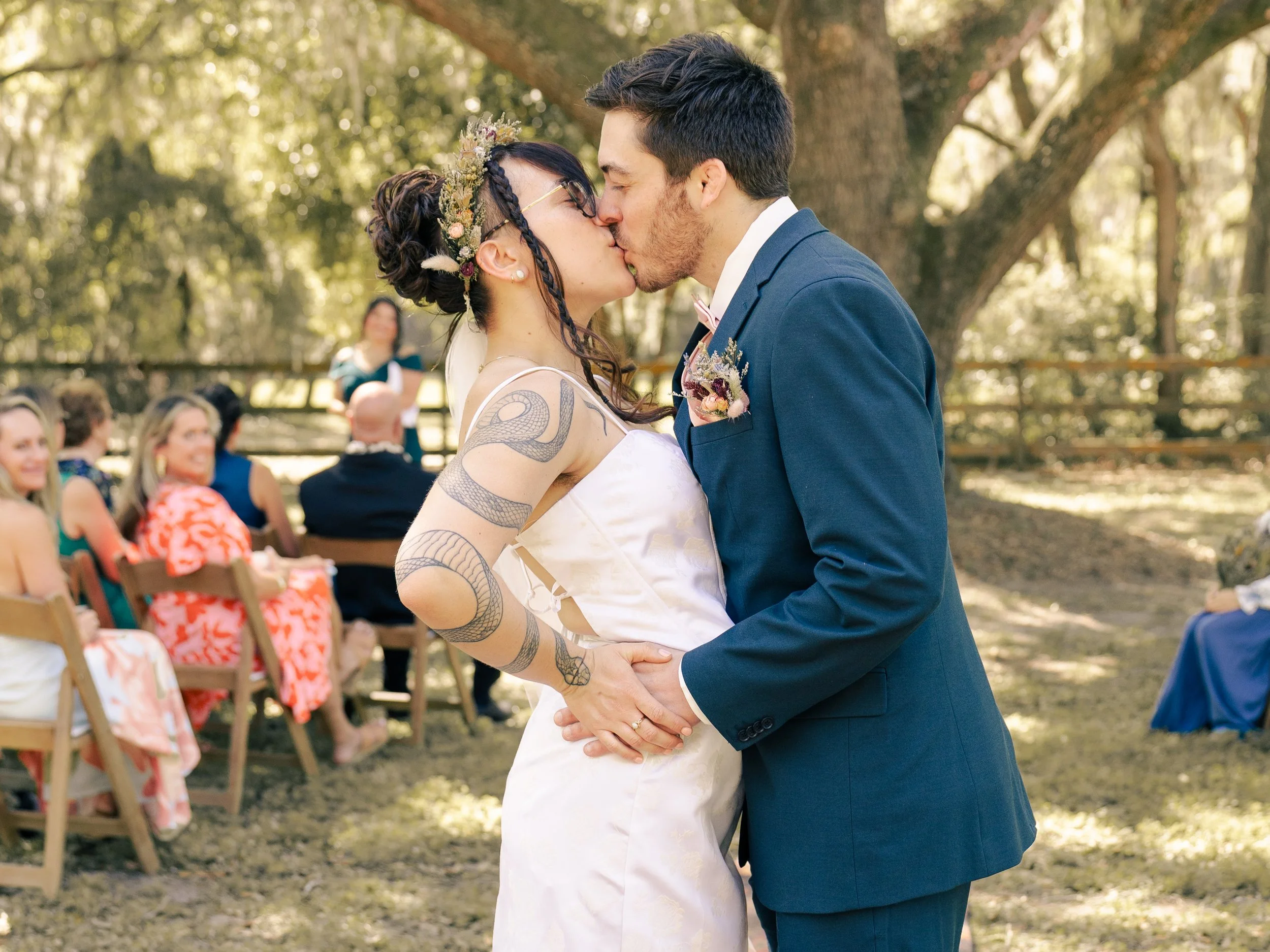 A couple is kissing during their outdoor wedding ceremony, with guests seated on chairs in the background. The woman has tattoos on her arm and is wearing a floral headpiece, and the man is dressed in a navy blue suit with a pink bow tie.