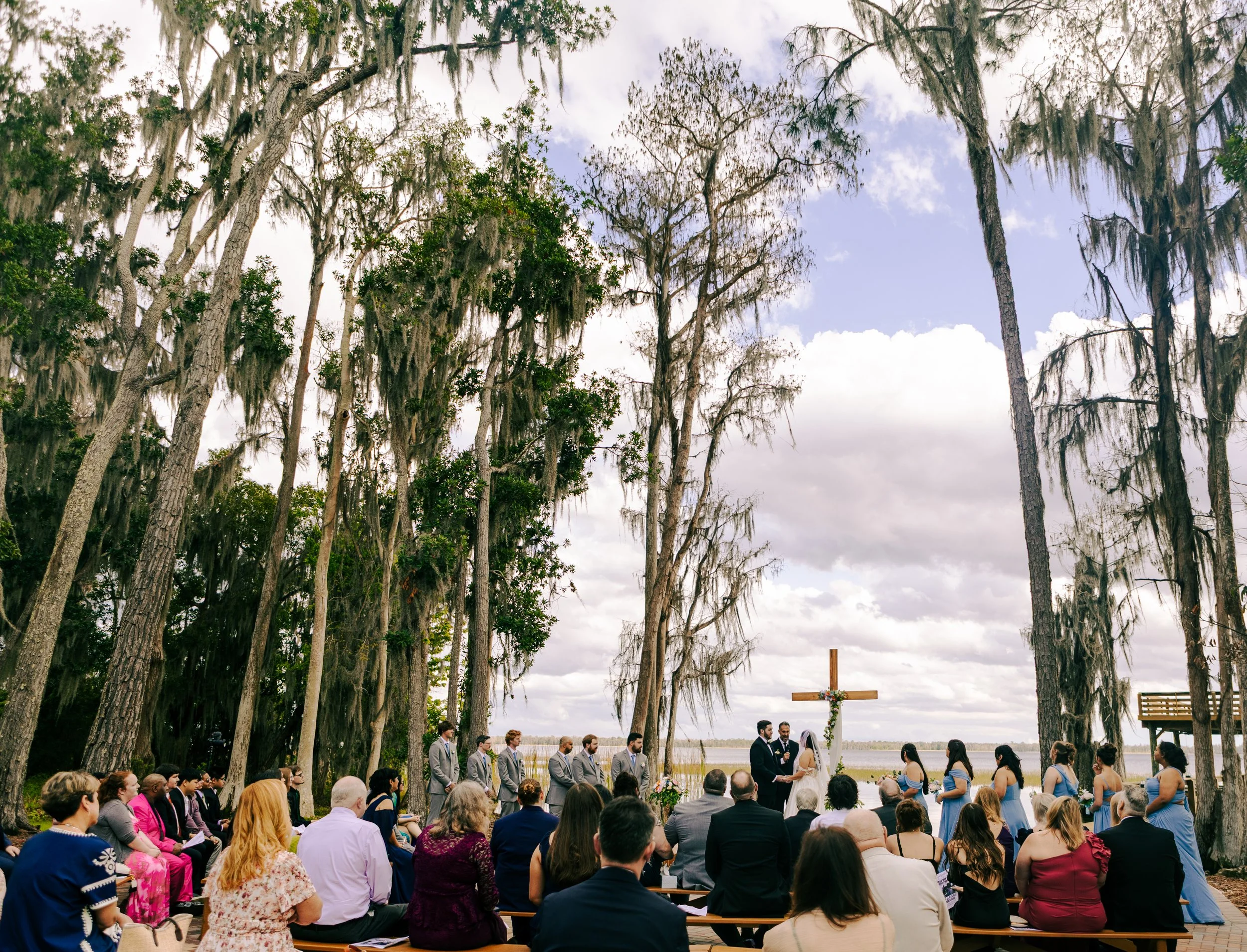 A large group of people attending a wedding ceremony outdoors, with some seated on benches and others standing. The ceremony is taking place under tall trees with Spanish moss hanging from them, near a body of water, with the wedding couple and offic