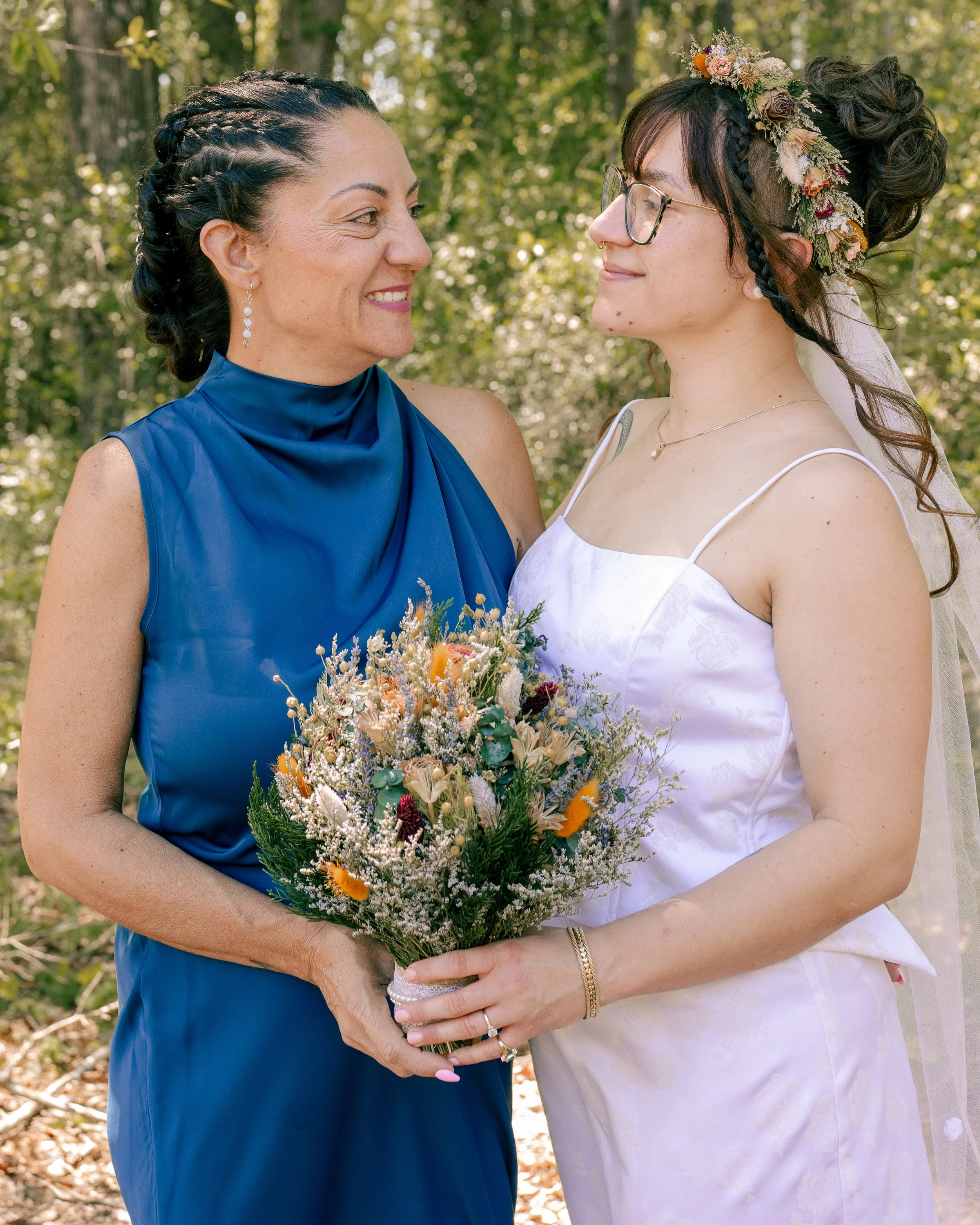 A woman in a white dress and a woman in a blue dress standing outdoors, holding a bouquet of flowers, facing each other and smiling.