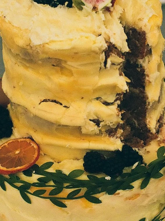 A partially sliced layered cake with vanilla frosting, decorated with blackberries, a blood orange slice, and green leaves.