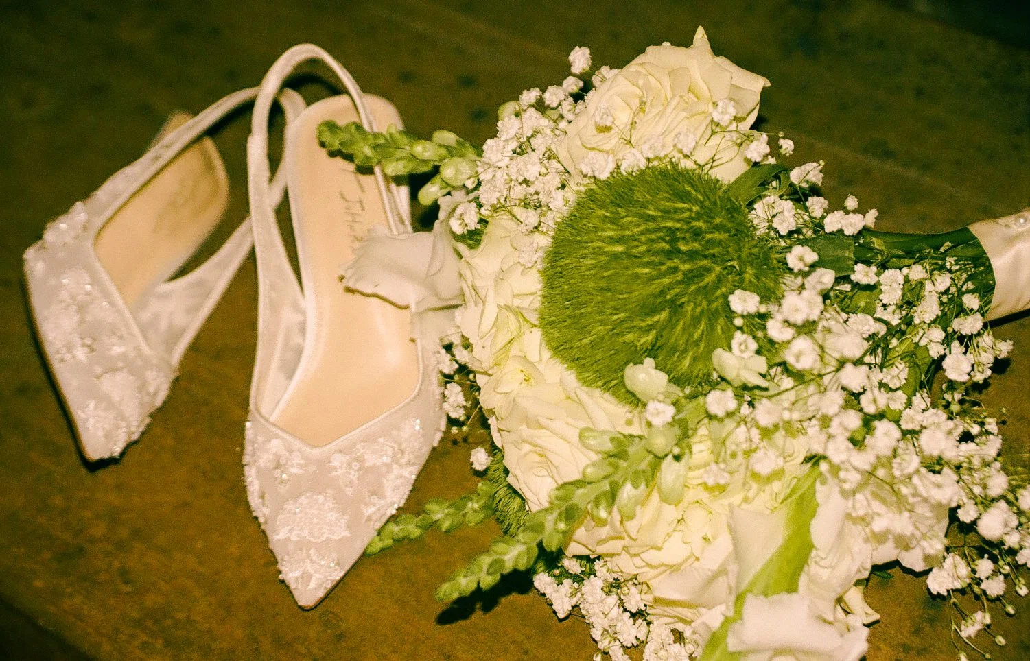 A bridal bouquet of white roses, green pom-pom flower, and baby's breath with white lace wedding shoes beside it on a dark surface.