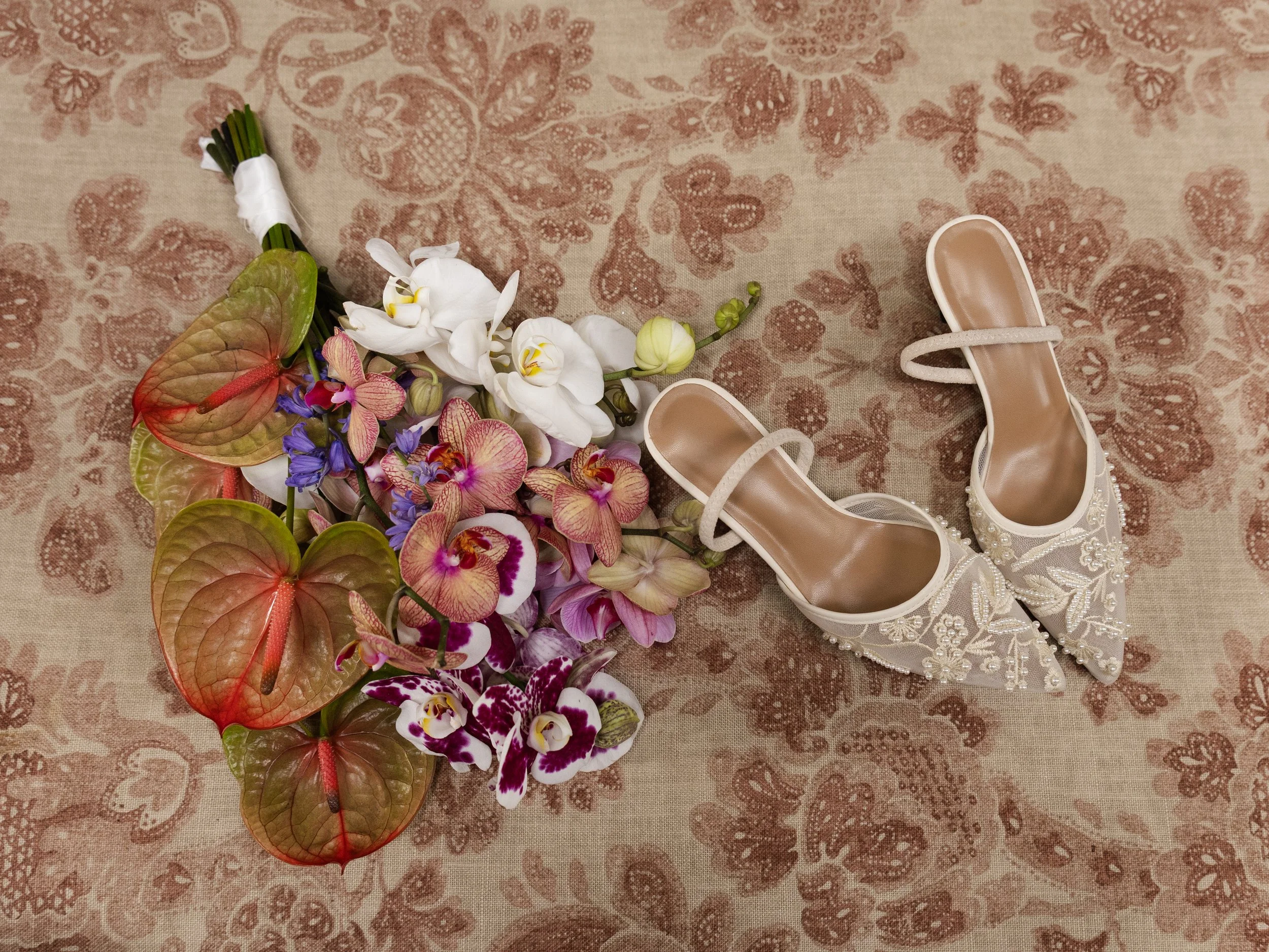 A pair of white lace bridal shoes with pearl embellishments, a bouquet of various flowers including orchids and anthuriums, all on a patterned fabric background.