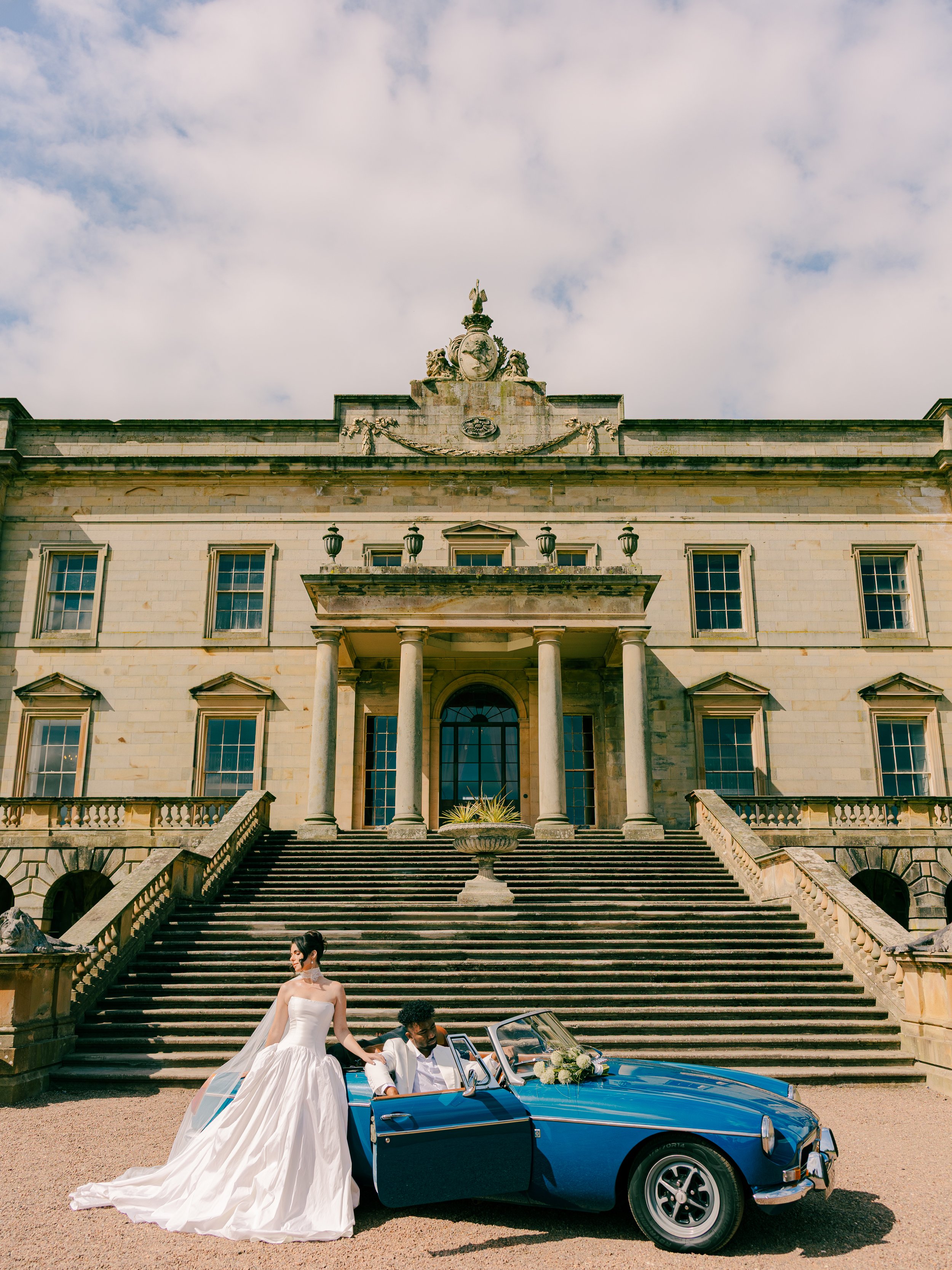 A bride and groom in a vintage blue convertible car in front of a grand historic building with steps and columns, wedding scene.