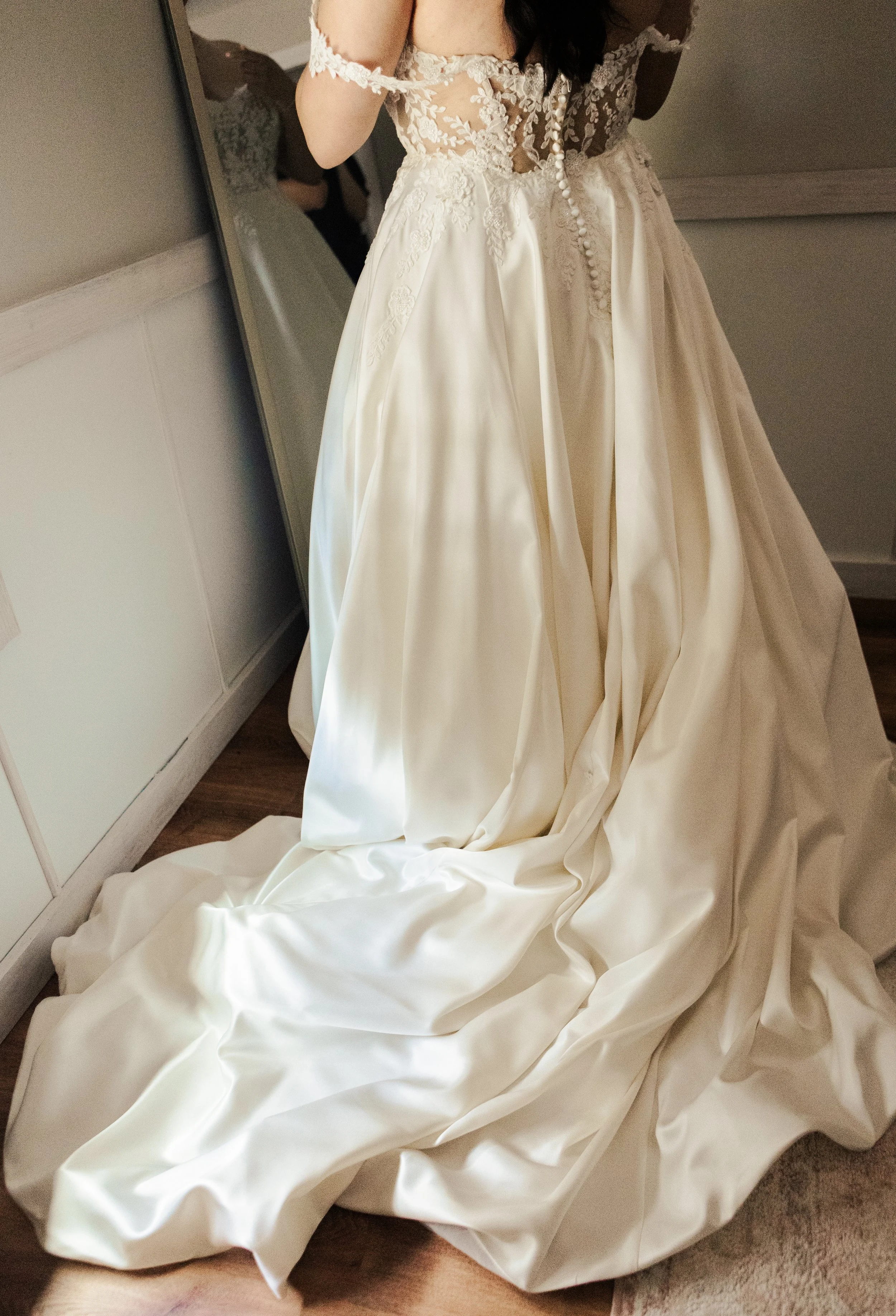 A woman trying on a wedding dress, taking a mirror selfie. The dress is ivory with lace detailing on the top and a full, flowing skirt that pools on the floor.