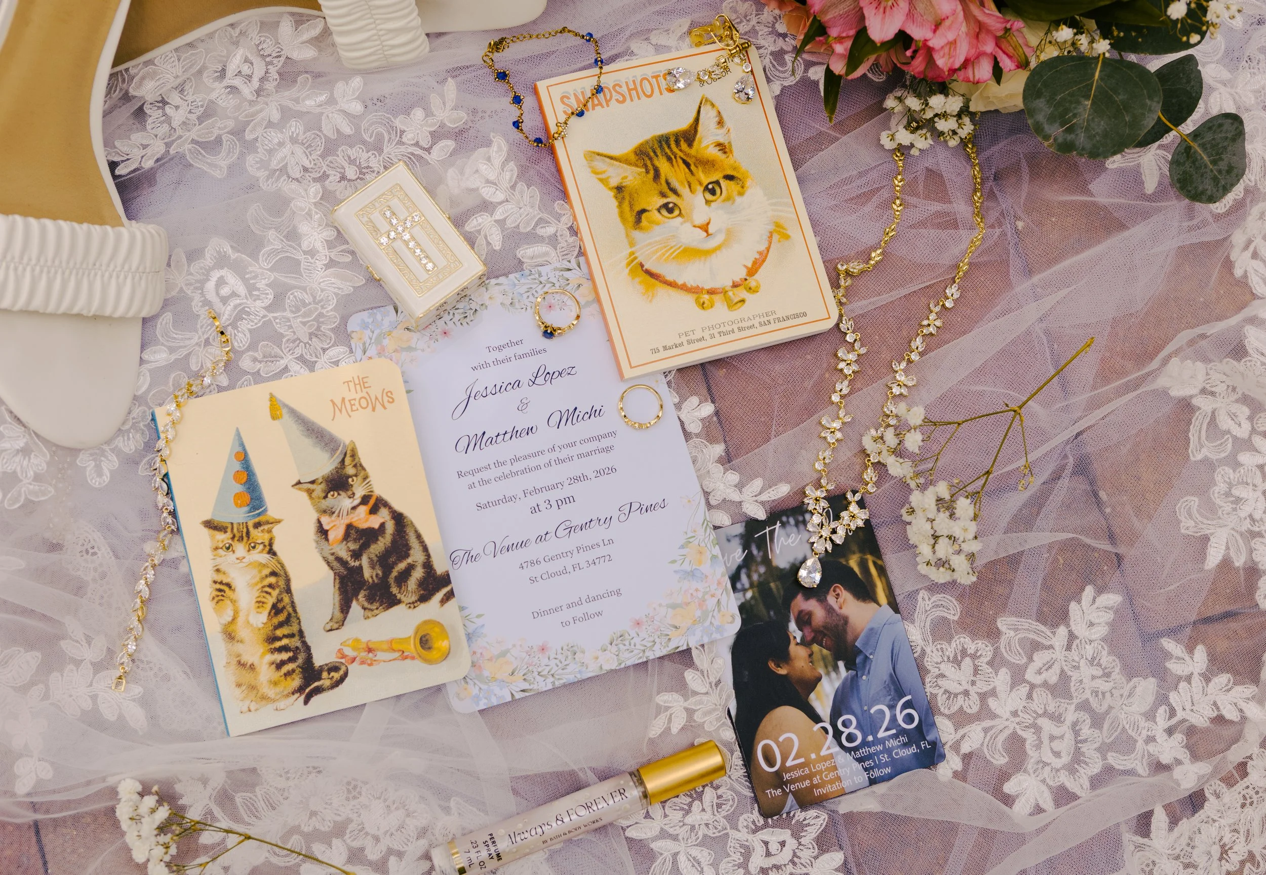 A wedding or celebration table decorated with jewelry, jewelry boxes, flowers, invitations, and photos. The jewelry includes necklaces, earrings, and rings, some with floral and diamond designs. The invitations feature a cat in a party hat and a coup