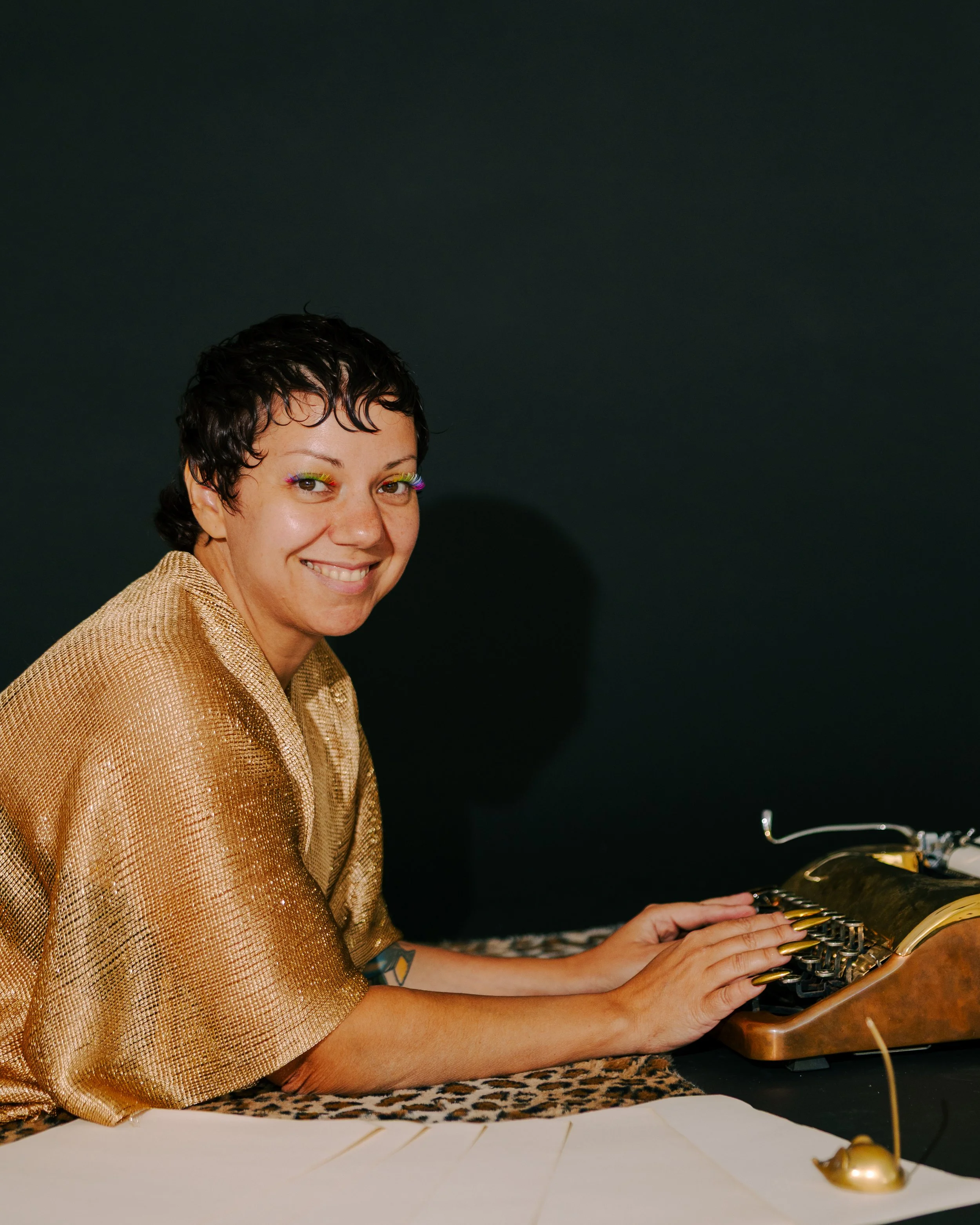 A smiling person with short dark hair, wearing colorful eye makeup and a shiny gold outfit, is playing a vintage typewriter. The scene is set against a dark background with a table covered in a leopard print cloth and some paper in the foreground.