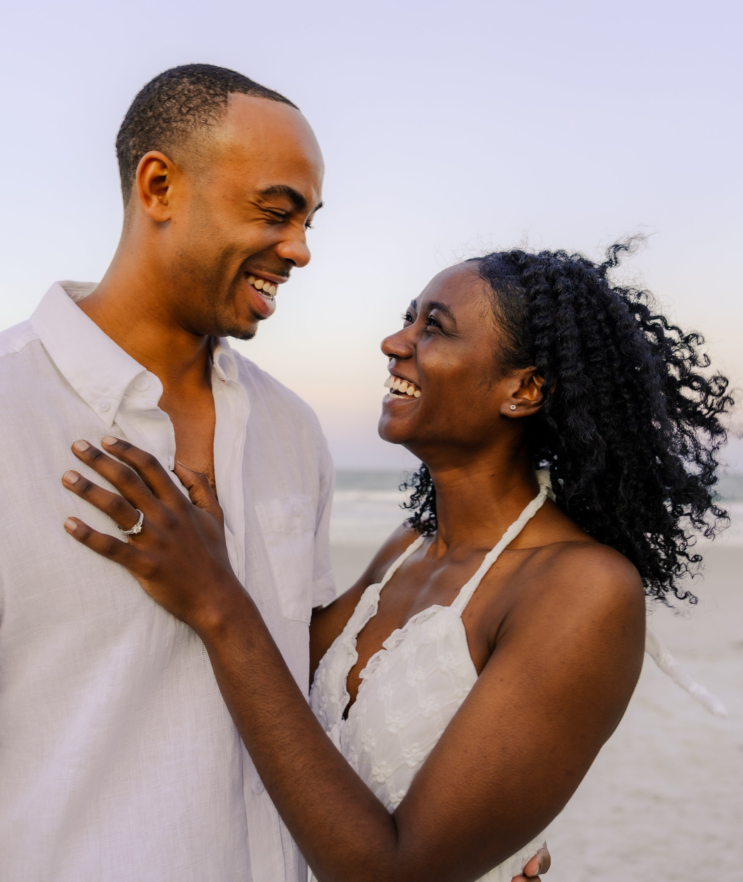 A smiling Black couple on the beach, looking at each other affectionately, with the ocean in the background.