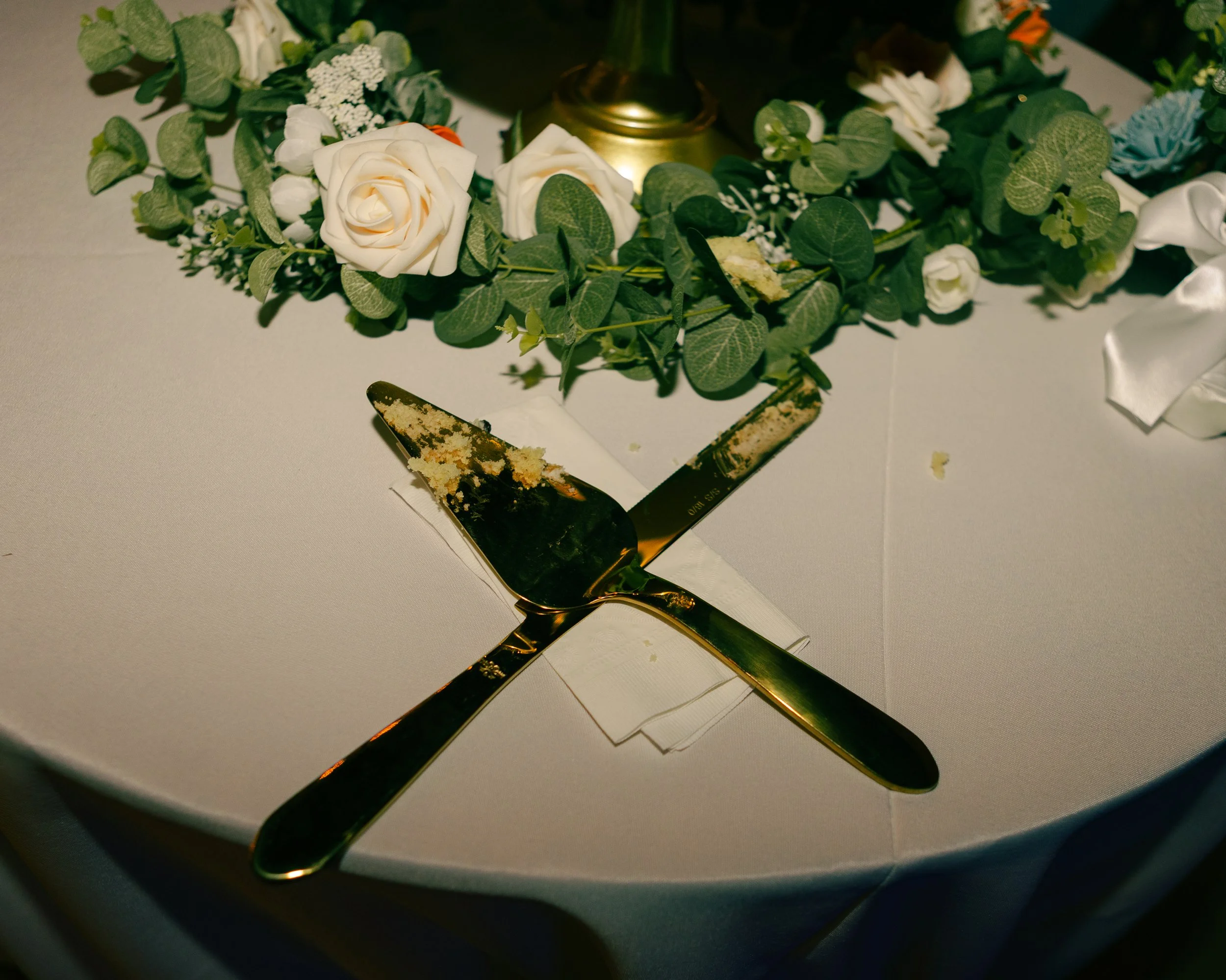 A floral arrangement with white roses, green leaves, and small white accents, surrounding a golden object on a white table, with a now-empty cake knife in the foreground.