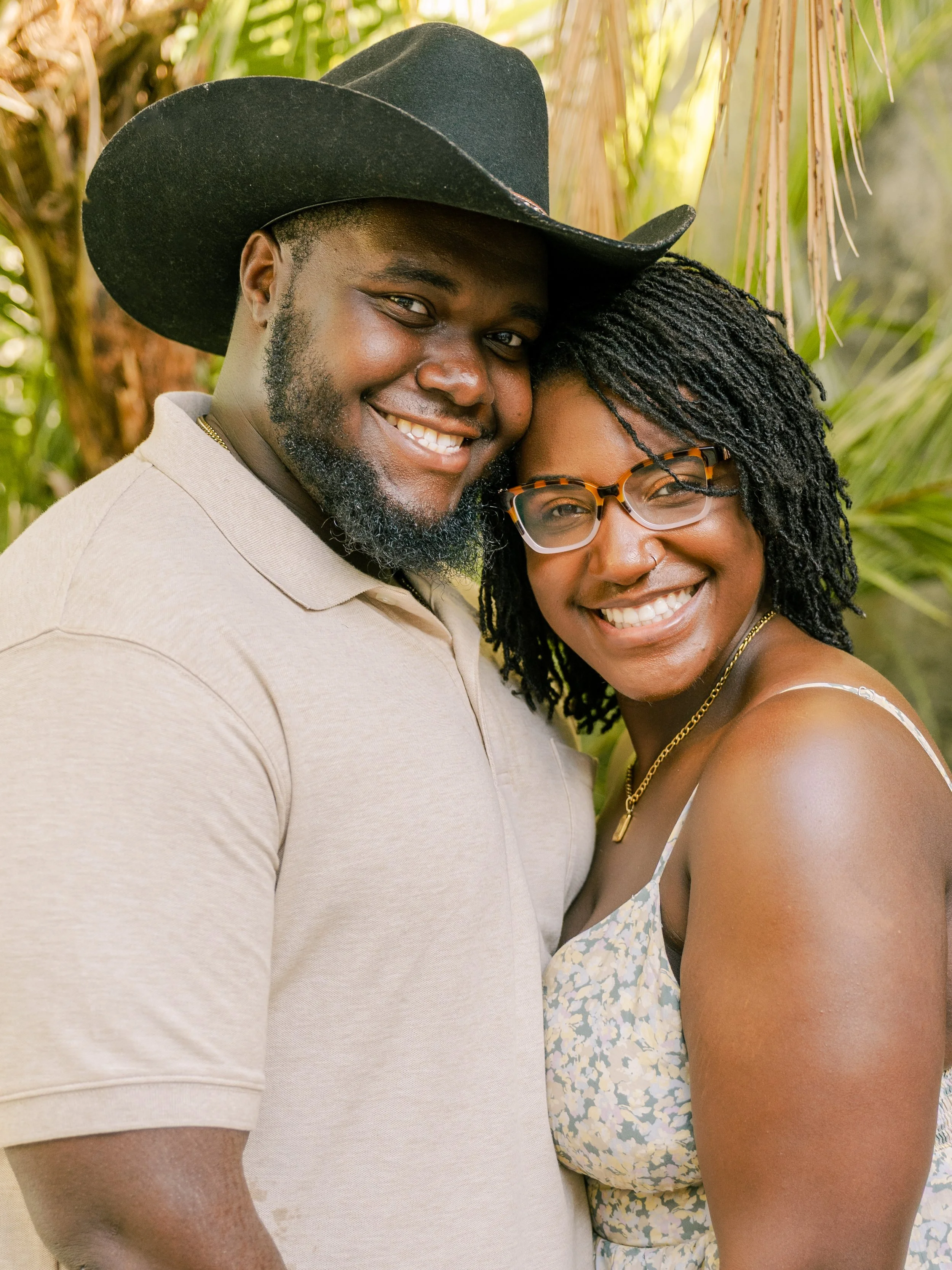 A smiling couple, a man with a cowboy hat and a woman with glasses, hugging outdoors with greenery in the background.