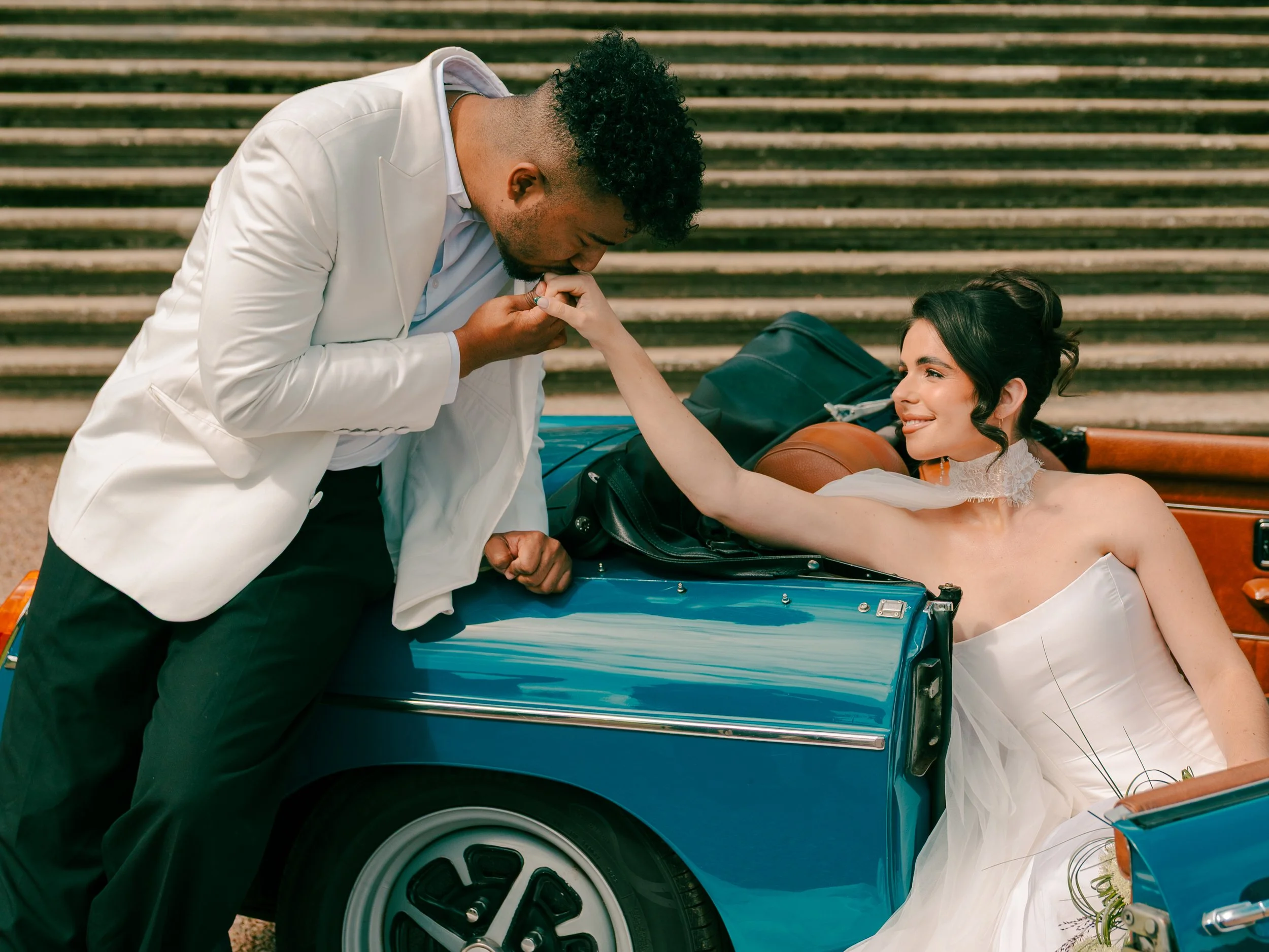 A man in a white suit kisses a woman in a white dress lying on a vintage blue car. The woman smiles, holding the man's hand, and is outdoors with steps in the background.