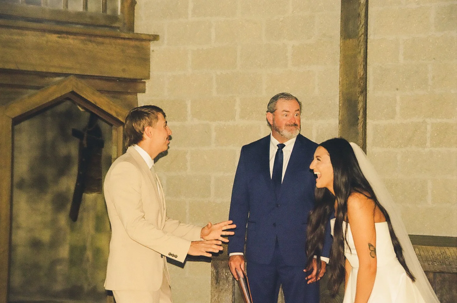 A bride and groom laughing and talking with a officiant inside a church during a wedding ceremony.