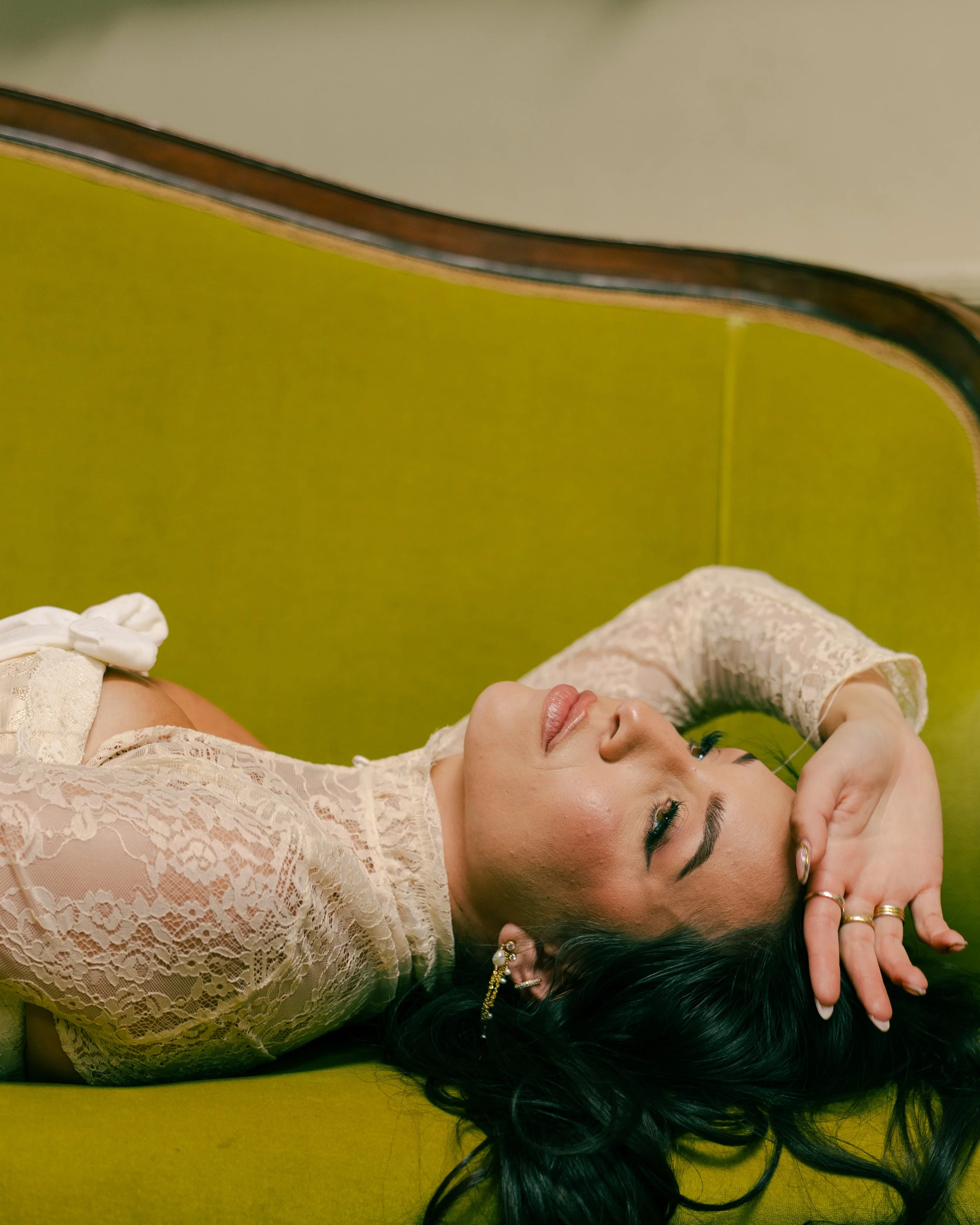 A woman with dark hair lying on her side on a yellow surface, wearing a cream-colored lace dress, with her eyes closed and one hand resting near her head.