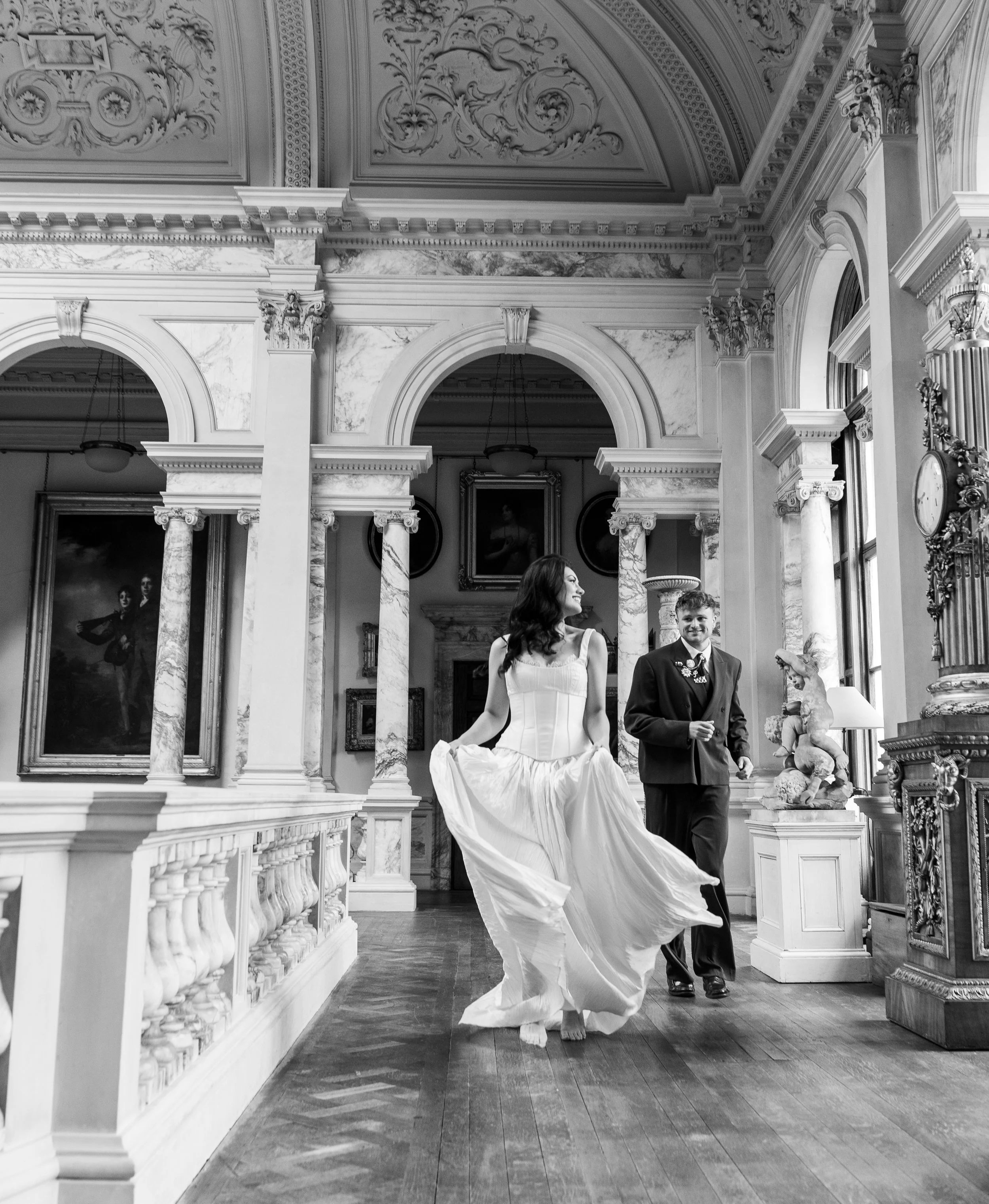 A black and white photograph of a woman in a wedding gown and a man in a suit dancing or posing in an elegant room with ornate architectural details, marble columns, and classical sculptures.
