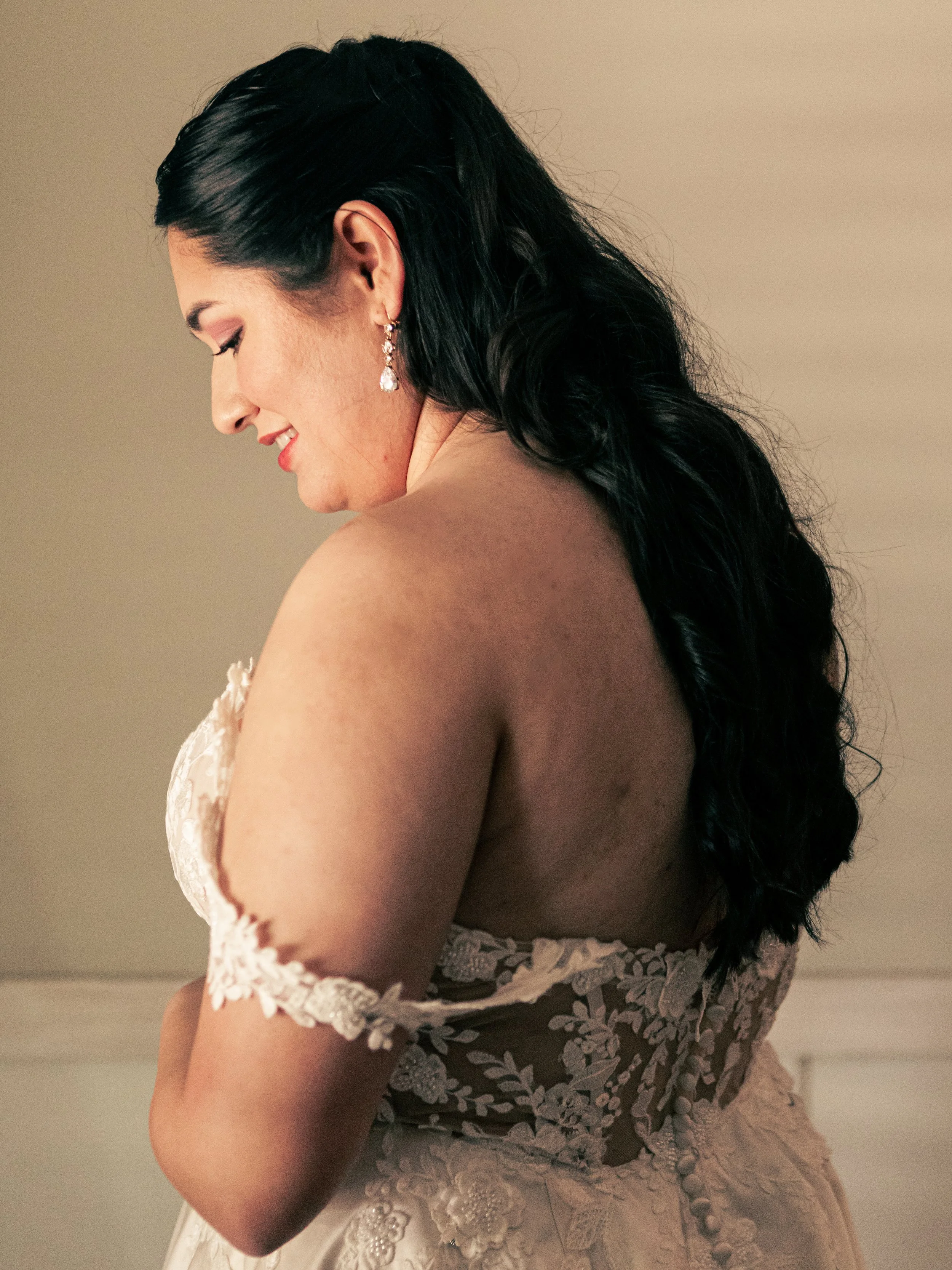 A woman with long black hair and drop earrings, wearing a lace dress with floral embroidery, looking down and smiling gently.