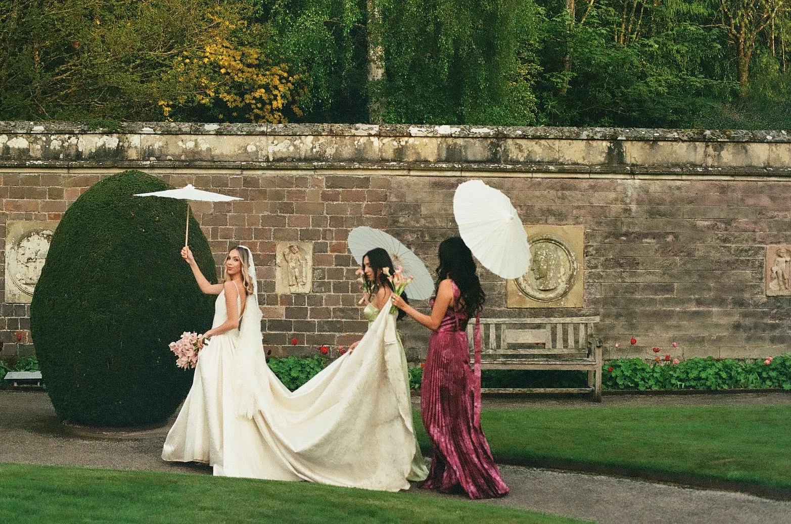 Three women in formal dresses holding umbrellas and flowers in a garden with a brick wall and statues.