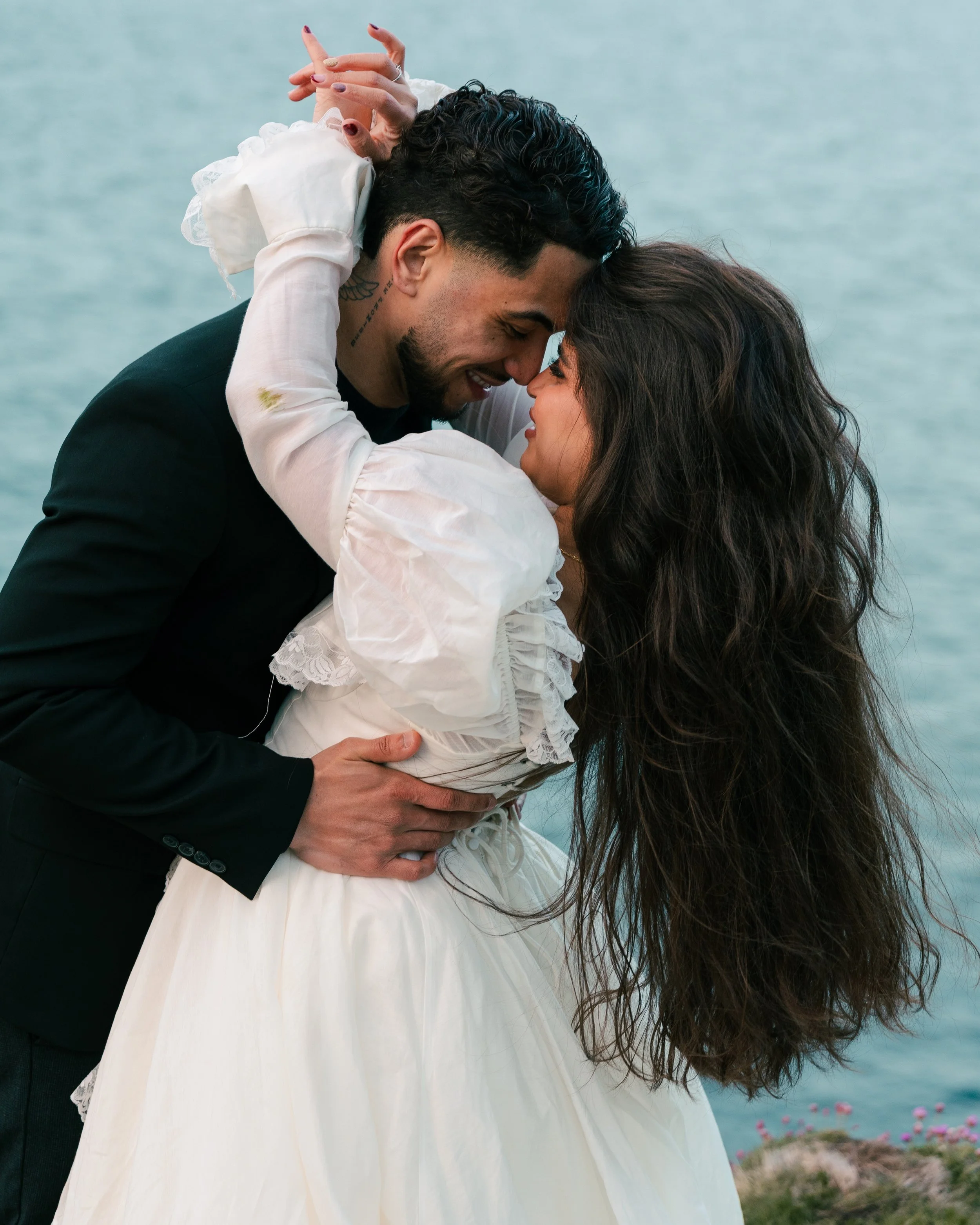 A couple embracing and touching foreheads by a body of water, with the man wearing a black suit and the woman in a white dress.