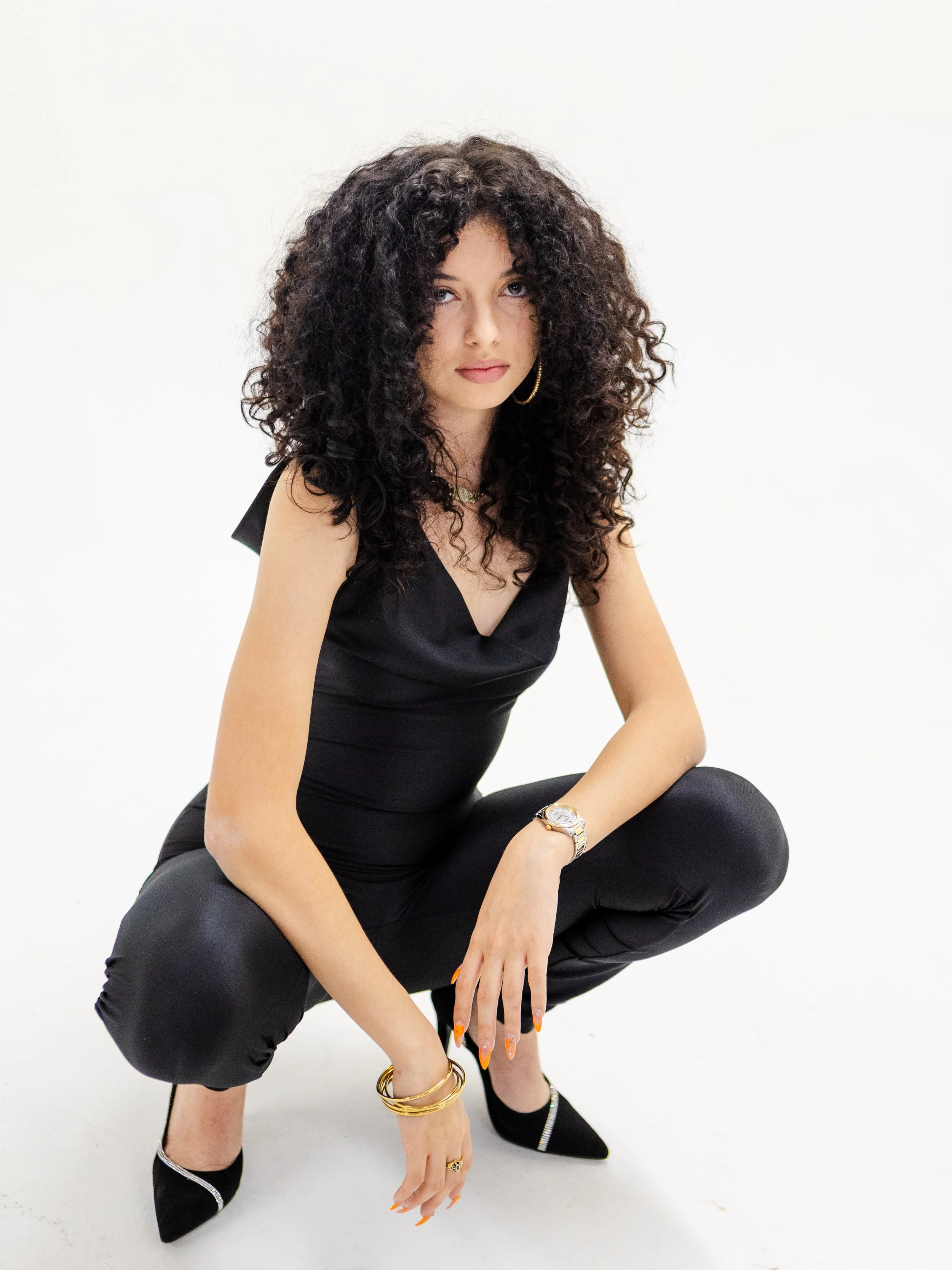 A woman with curly hair looks at the camera wearing a black jumpsuit