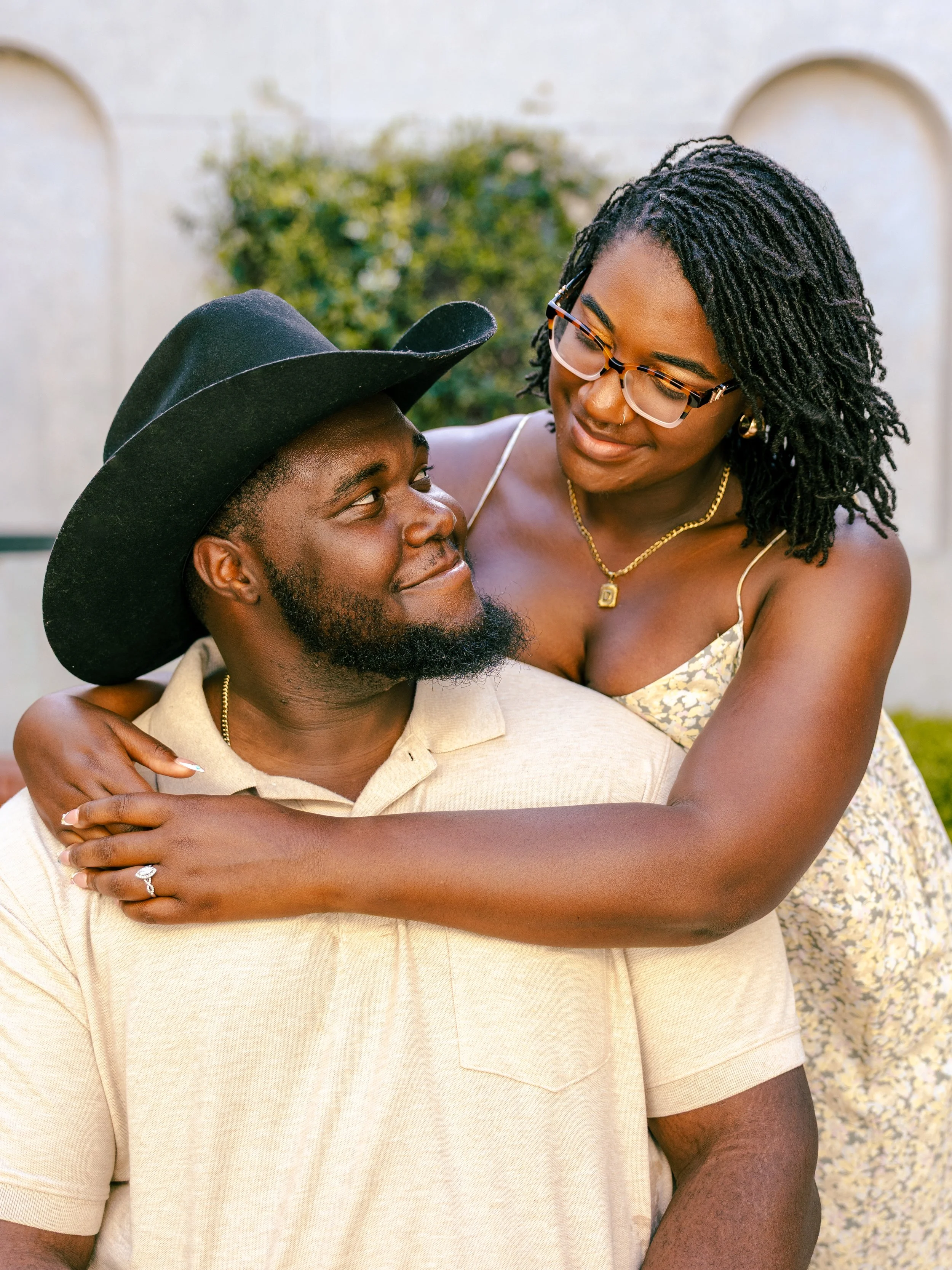 A man and woman sharing a hug outdoors, smiling at each other. The man has a beard and is wearing a beige shirt and a black hat. The woman has black, curly hair, glasses, and is wearing a floral dress with jewelry.